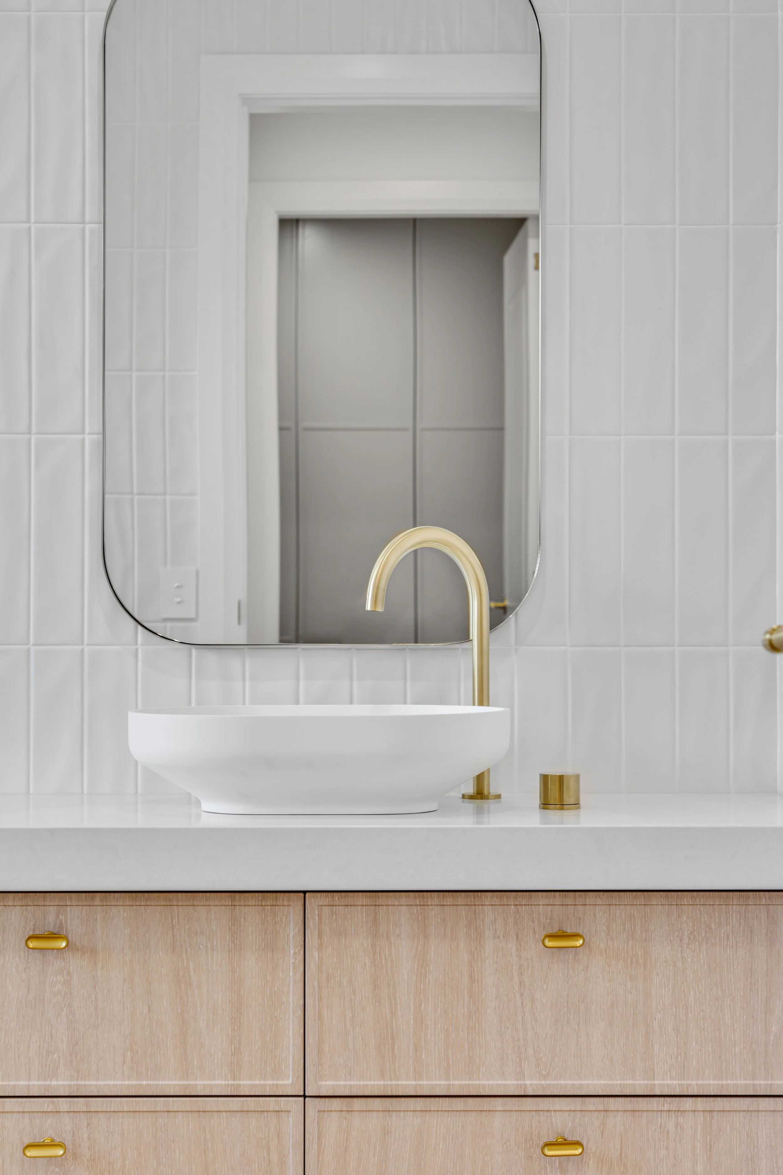 A modern bathroom vanity with a white rectangular sink, gold faucet, and beige cabinet with gold handles. A large mirror is mounted on a tiled wall, with a reflection of a hallway or another room.