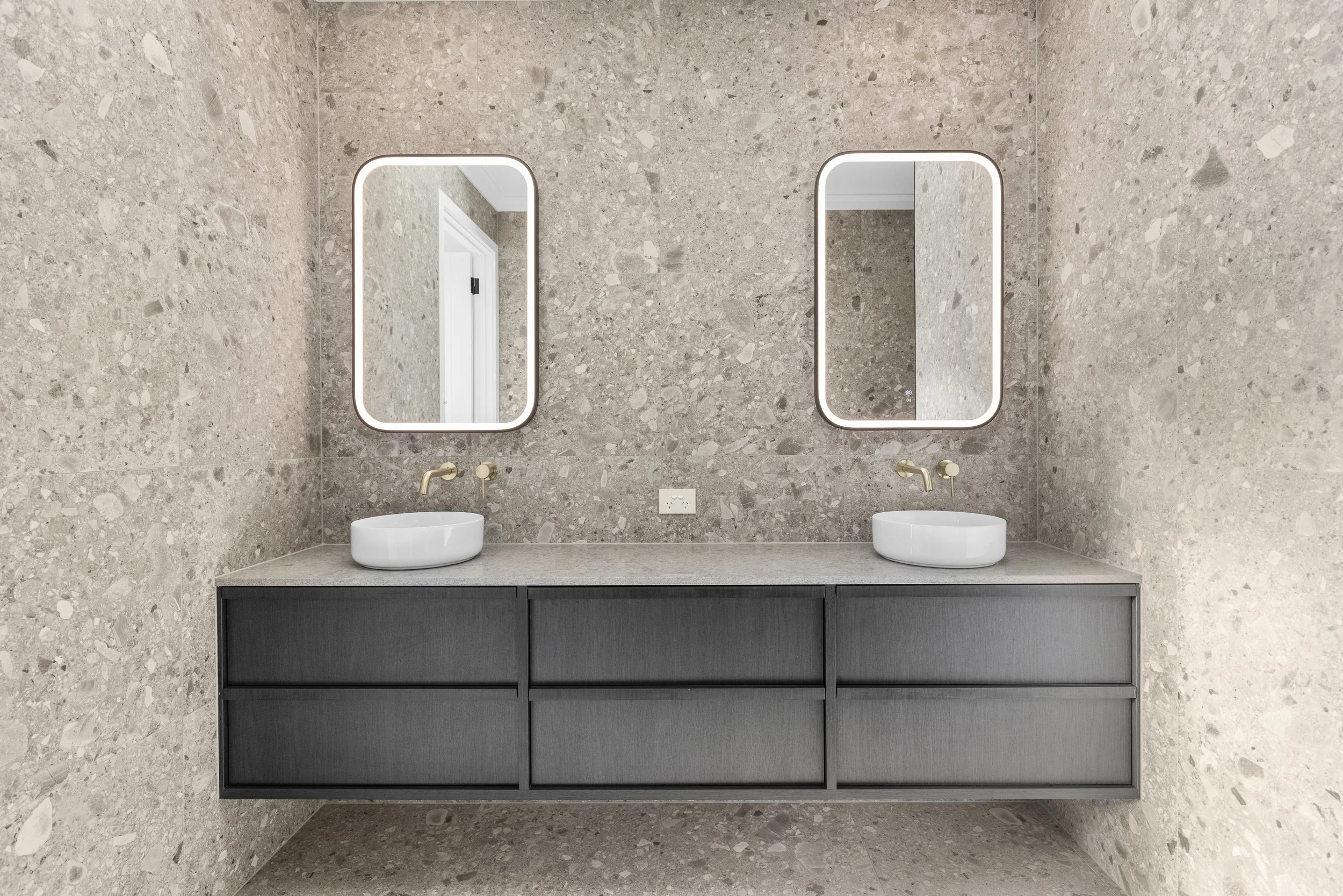 A modern bathroom with a beige stone wall, two oval mirrors with LED lighting, and two white vessel sinks on a black vanity.