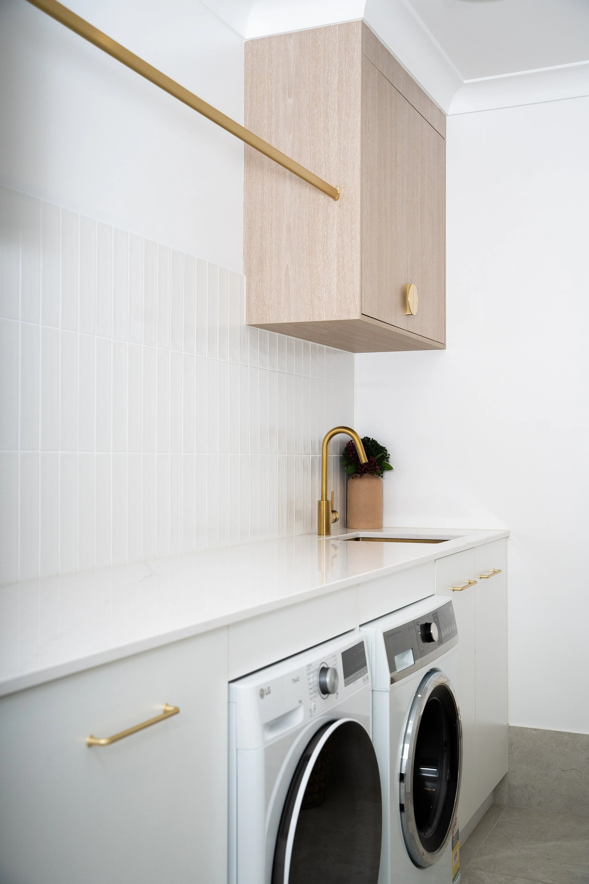 A laundry room with white washer and dryer side by side under a white countertop. There is a beige cabinet above the appliances and a gold faucet with a potted plant nearby.