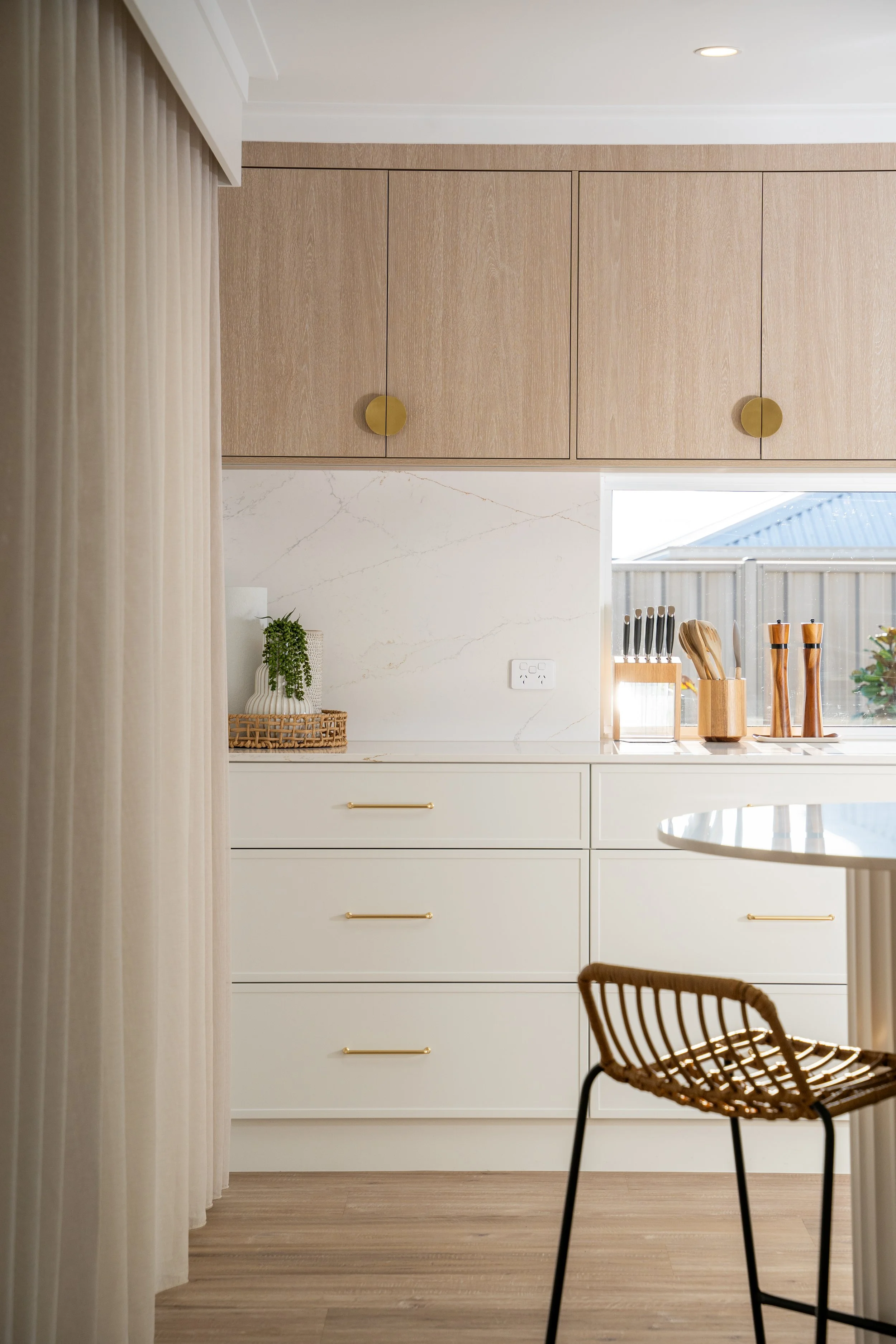 Modern kitchen with beige upper cabinets, white lower drawers, marble backsplash, and a round wooden chair in the foreground.