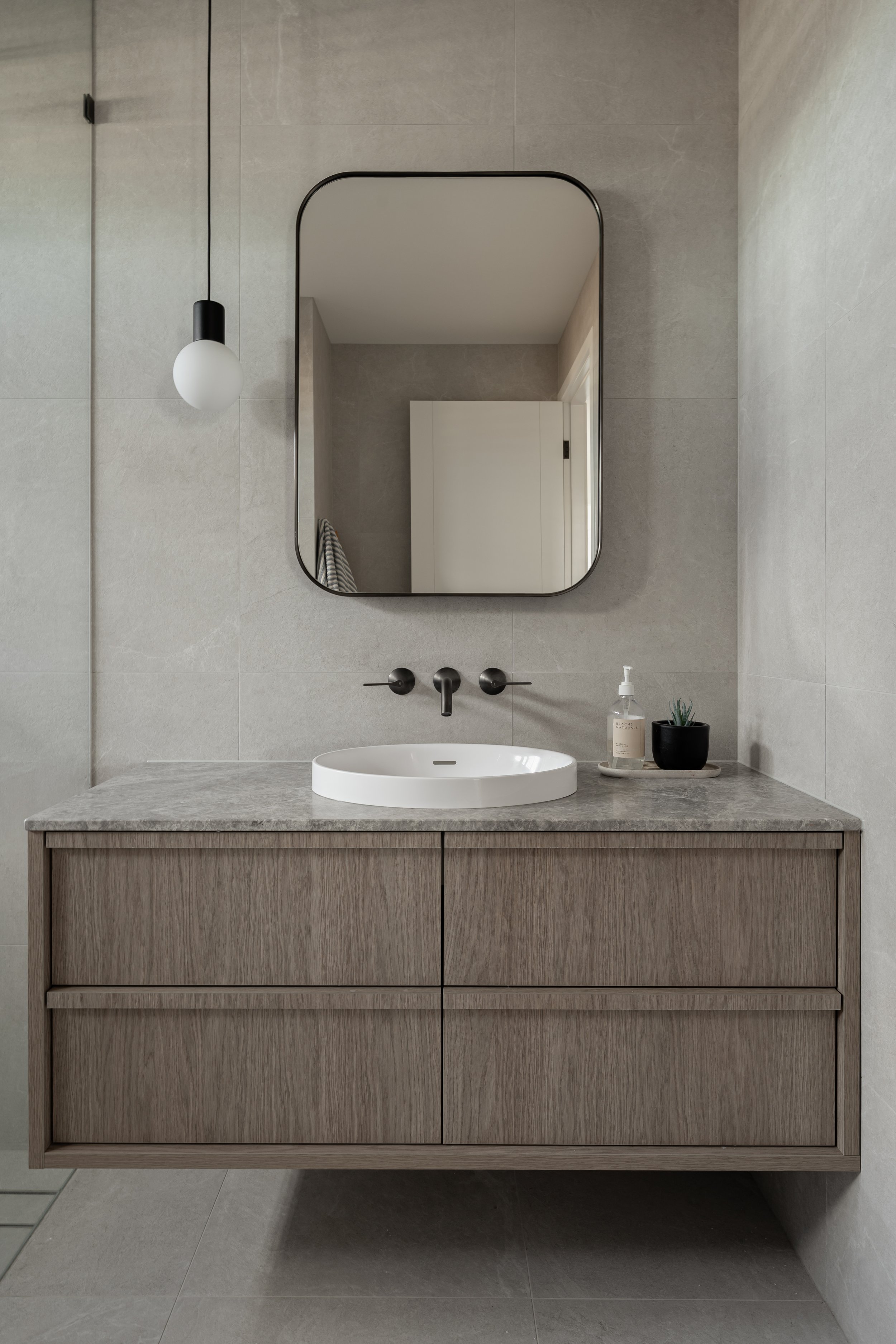 A modern bathroom vanity with a sink, mirror, hanging pendant light, and a soap dispenser, set against a wall with large light gray tiles.