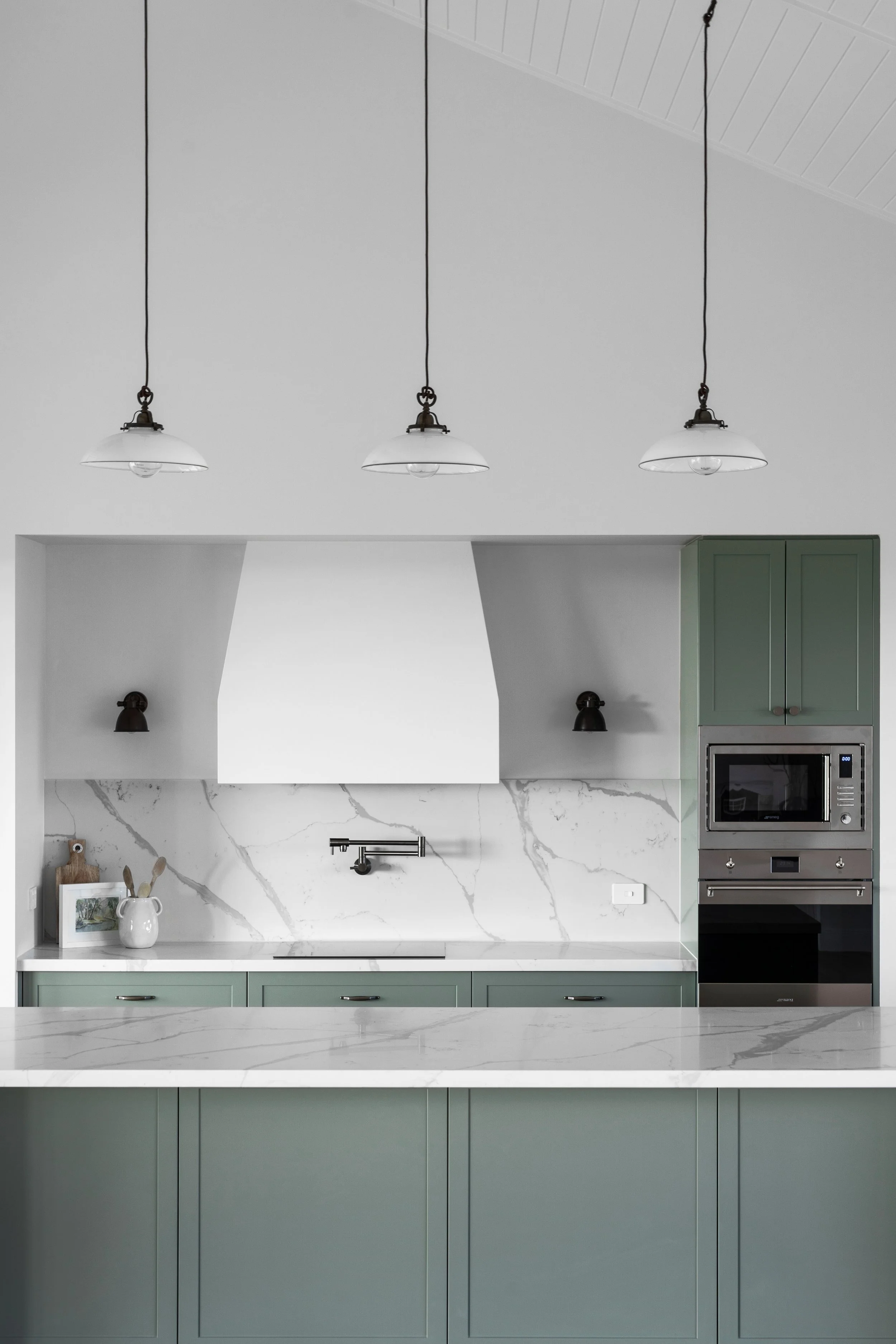 Modern kitchen with green cabinets, marble countertop and backsplash, white range hood, built-in microwave, and hanging pendant lights.