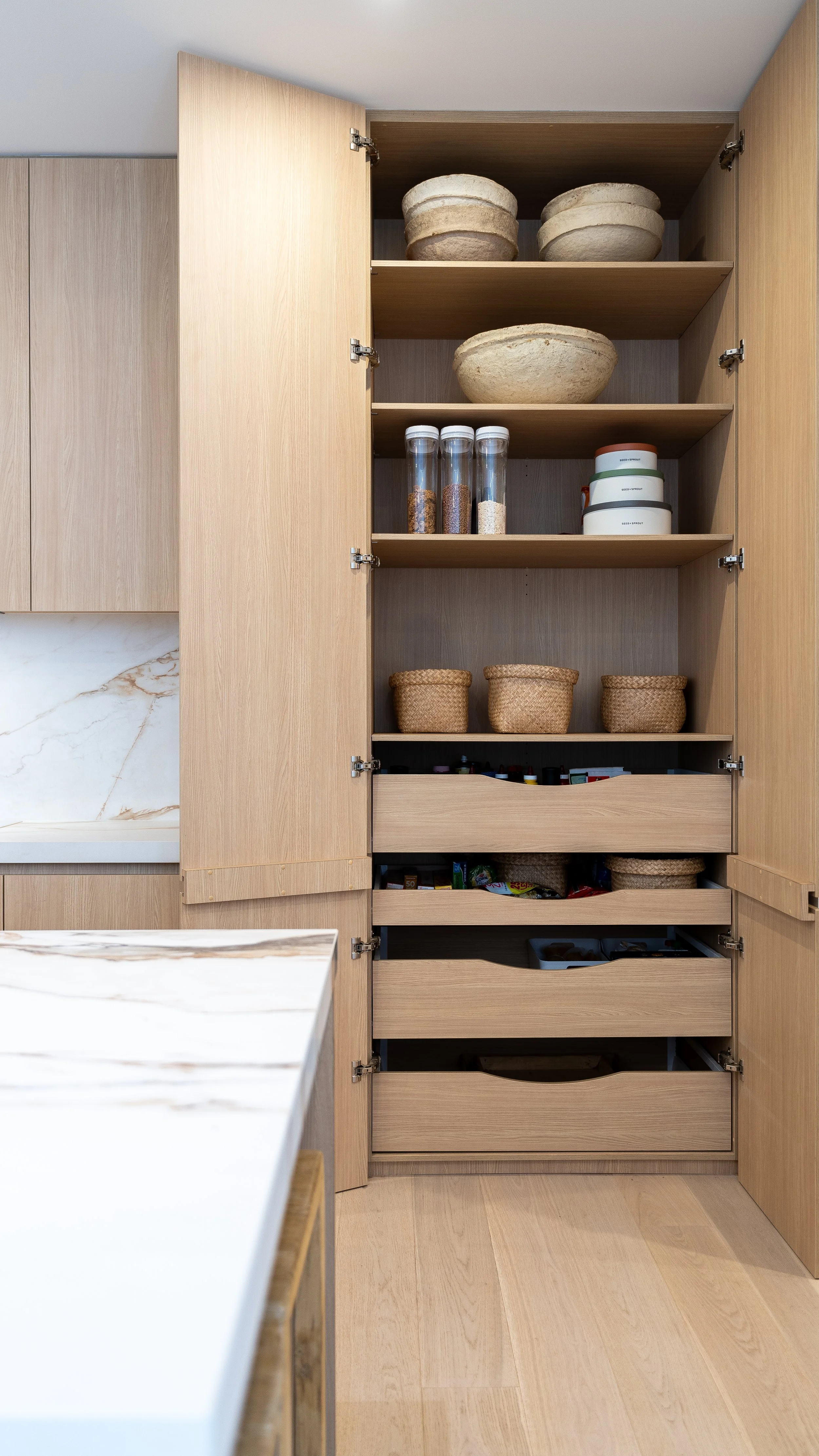 Open kitchen cabinet with multiple shelves storing containers, bowls, and jars, made of light wood.