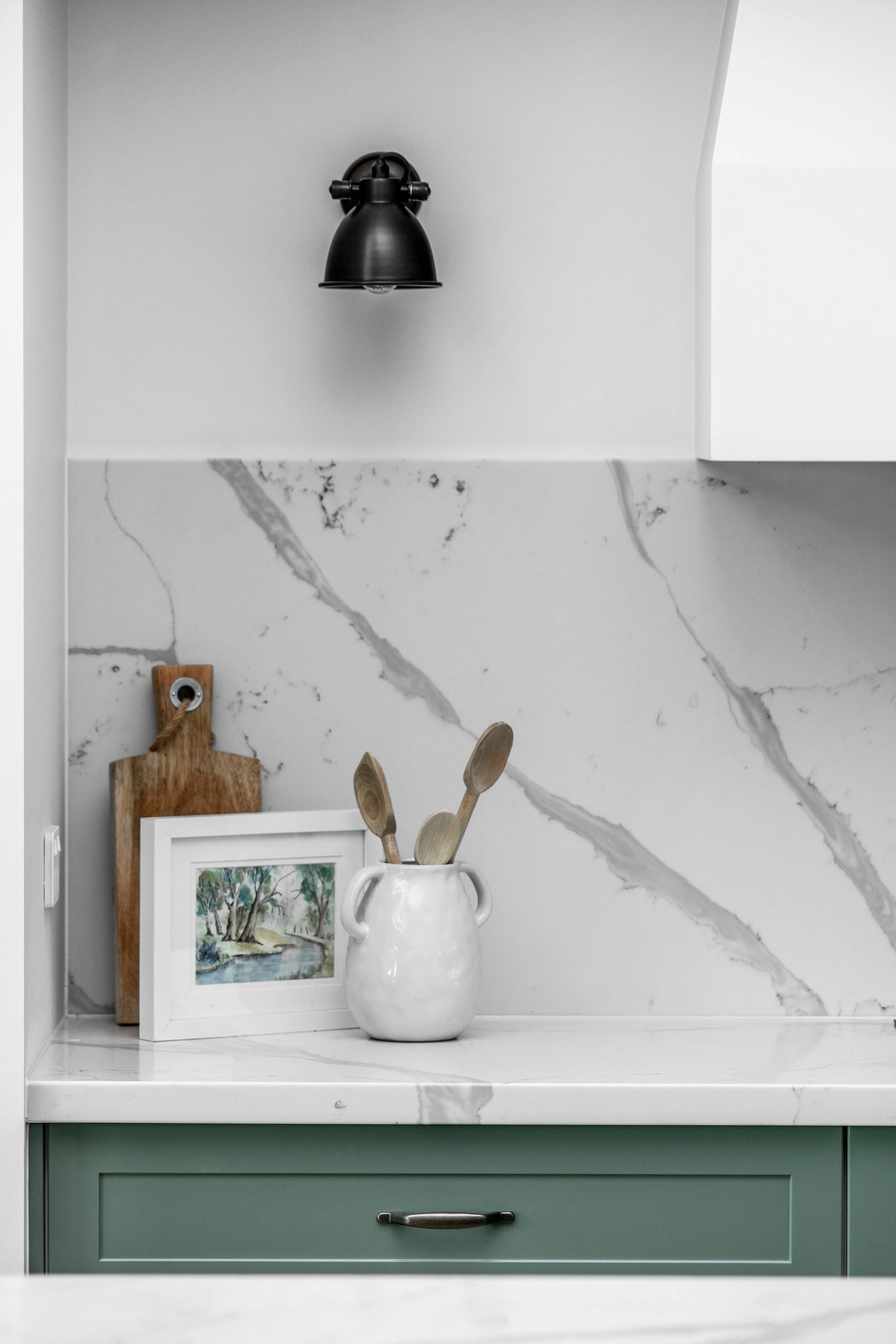A kitchen countertop with a white vase holding wooden spoons, a framed watercolor painting of trees and a river, a cutting board, and a marble backsplash with gray veining.