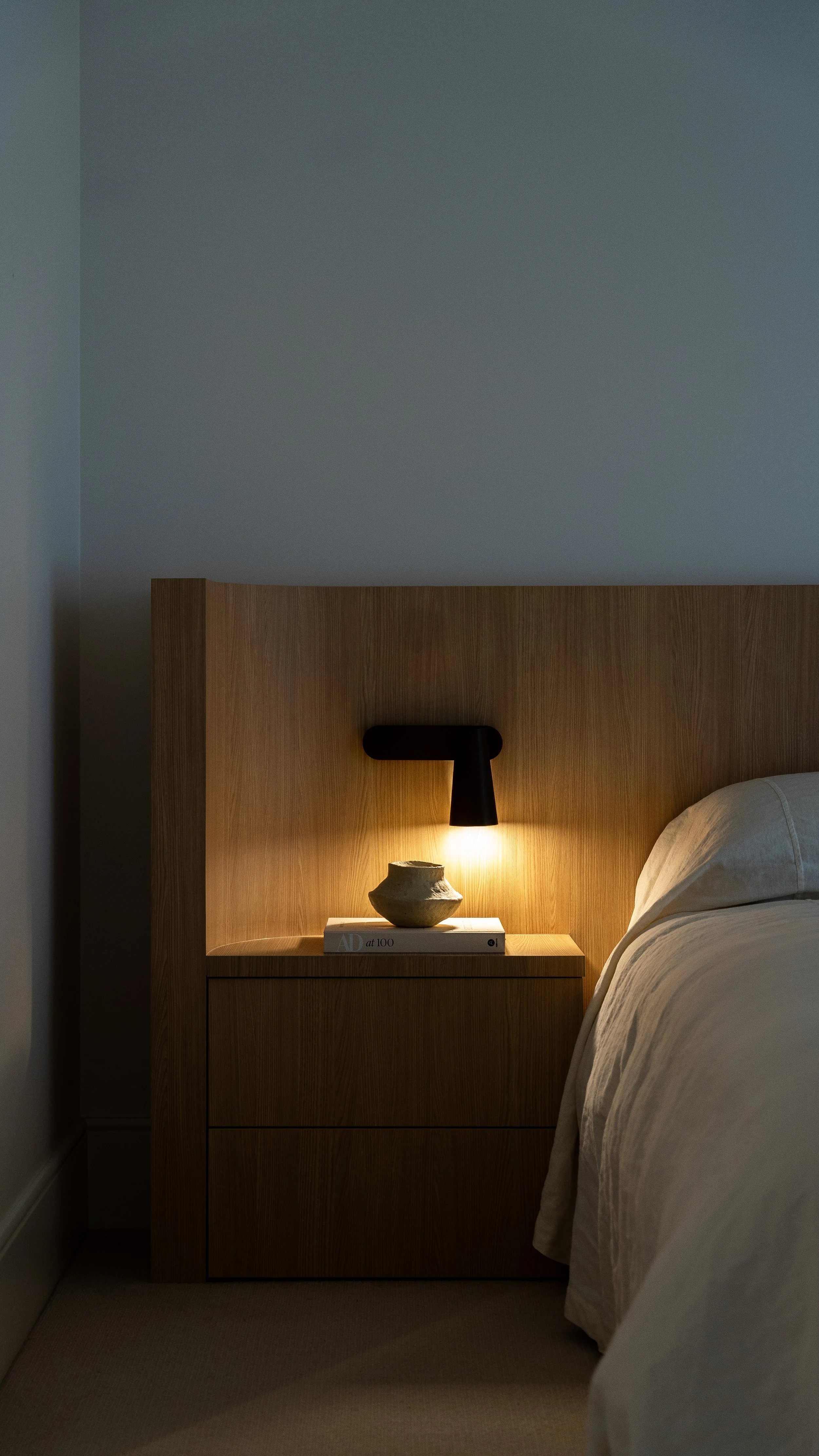 A bedroom nook with a wooden headboard, a black wall-mounted bedside lamp, a beige vase, a book titled 'AD at 100', and a beige nightstand.