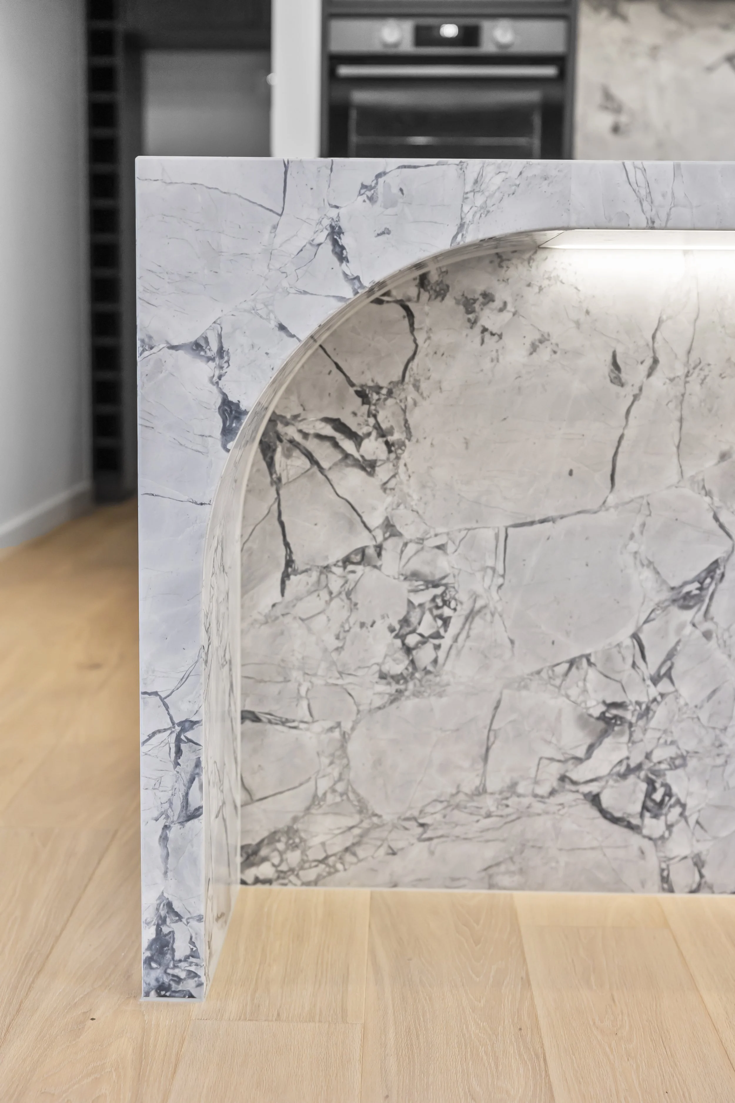 Close-up of white marble with gray veining on a rectangular surface and a rounded cutout, set on a wooden floor with a black appliance in the background.