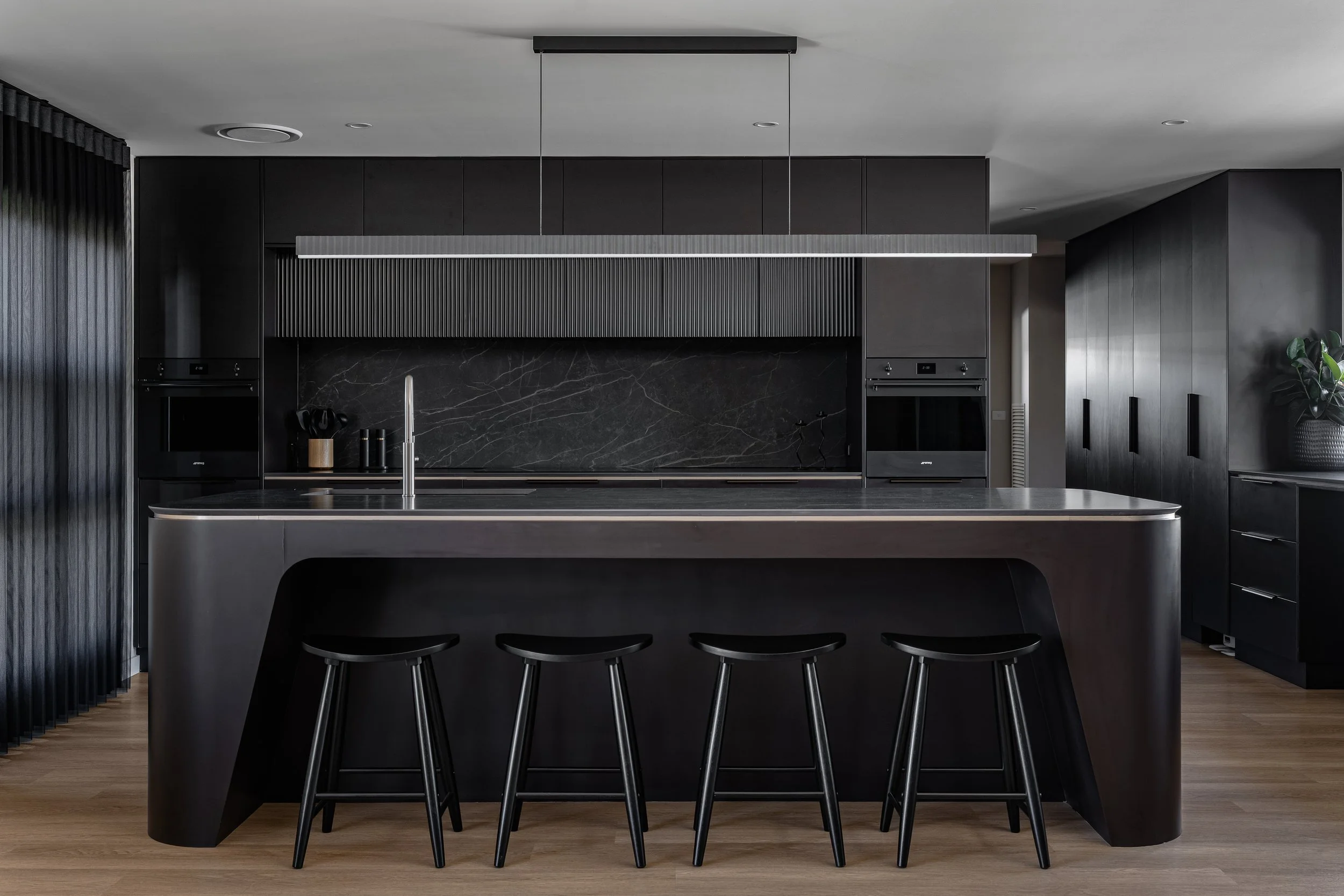 Modern black kitchen with a large island, black cabinetry, and wood flooring. The island has a black countertop with a sleek metallic faucet and four black stools underneath. The background features black cabinets, a black marble backsplash, and buil