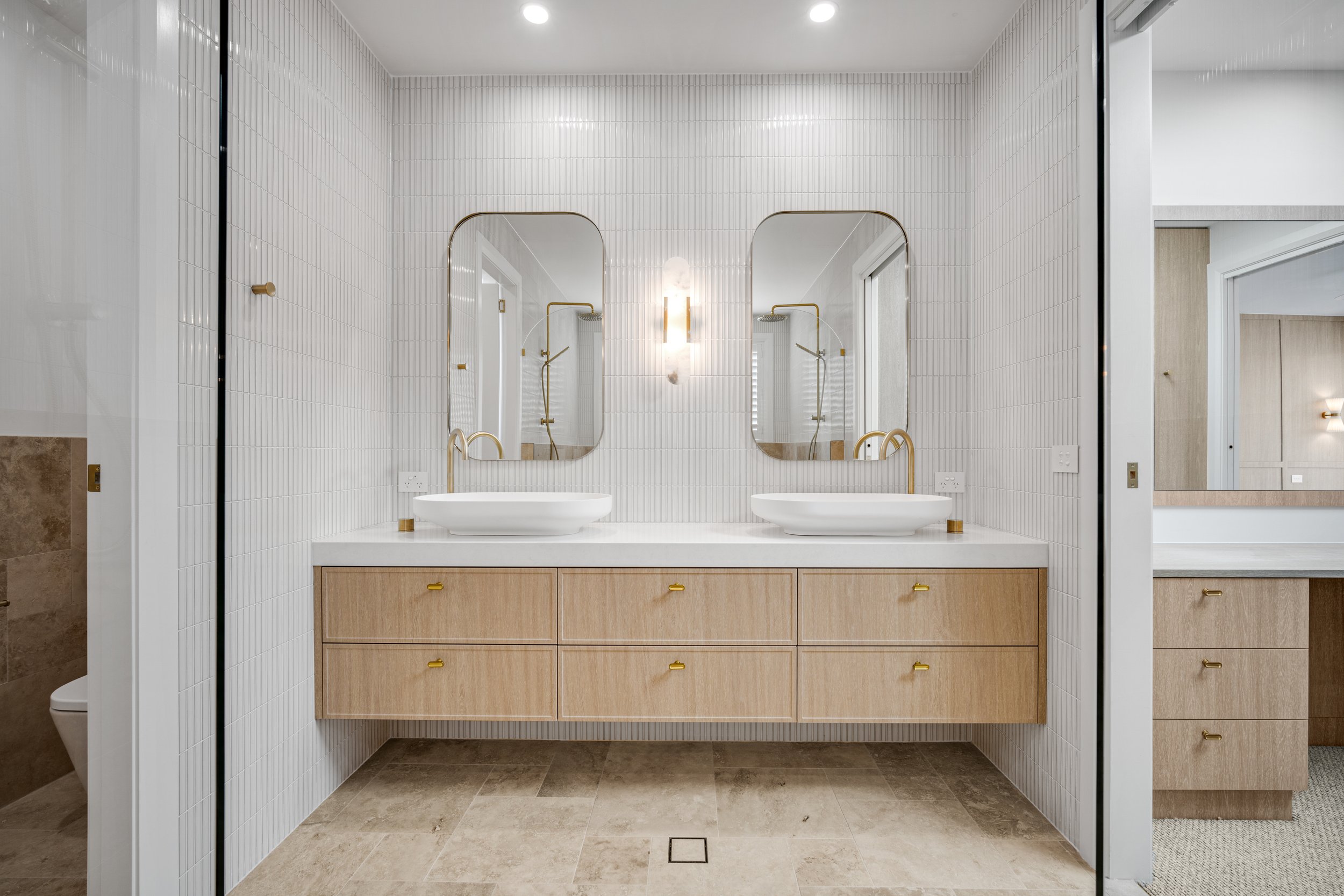 Modern bathroom with two white vessel sinks, gold faucets, large mirrors, and wood vanity with multiple drawers, white tiled walls, and beige tile flooring.
