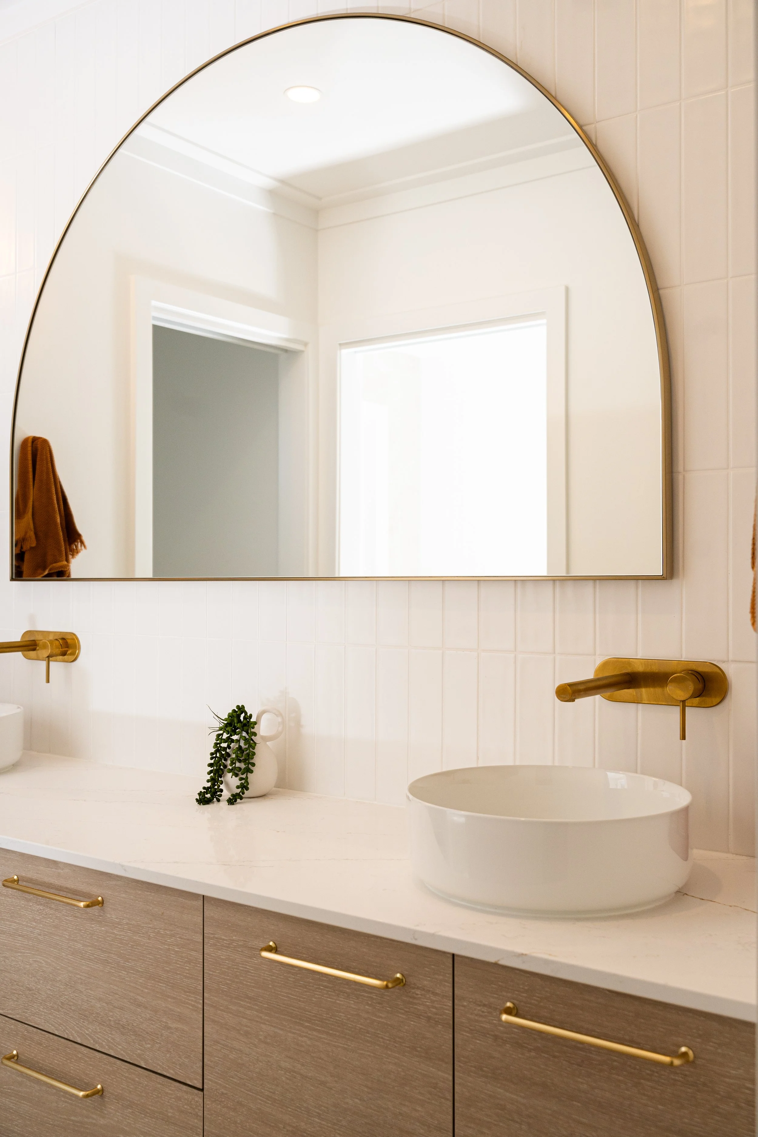 Modern bathroom vanity with a large mirror, white countertop, a vessel sink, gold fixtures, and a small green plant in a white vase.
