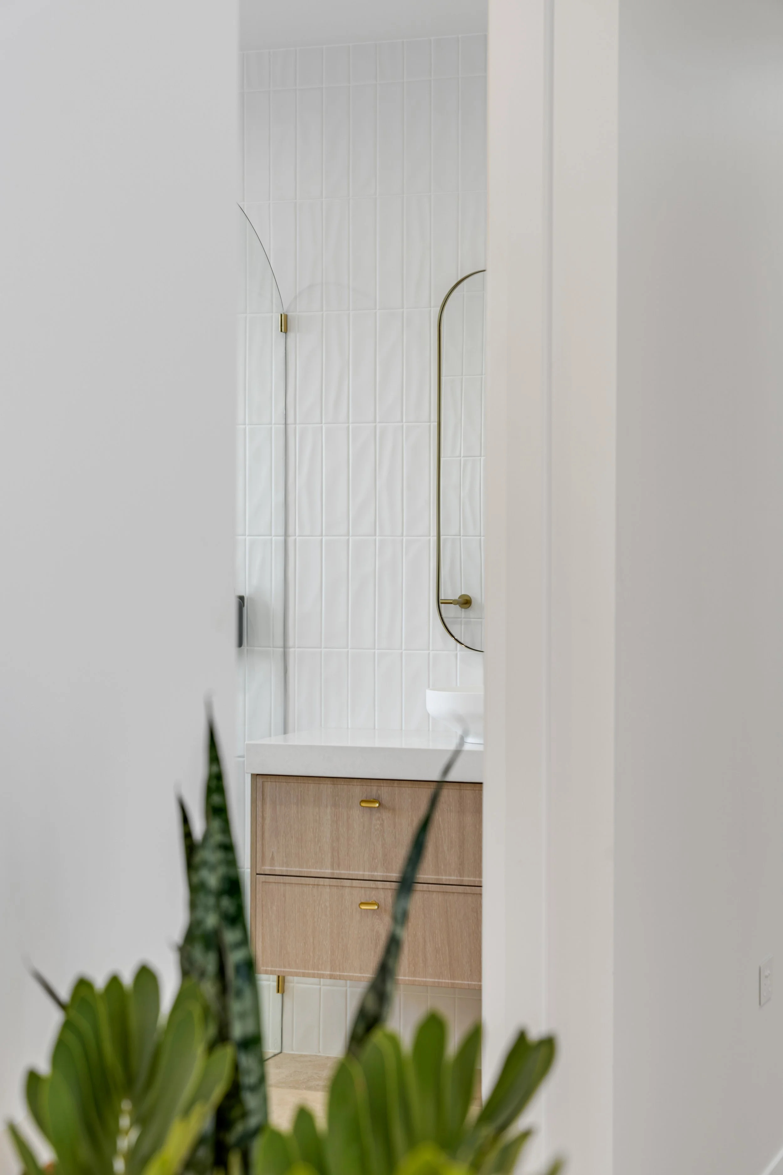 View of a bathroom through an opening, showing a wall with white tiles, a wooden cabinet with yellow handles, a white sink, and a decorative leaf plant in the foreground.