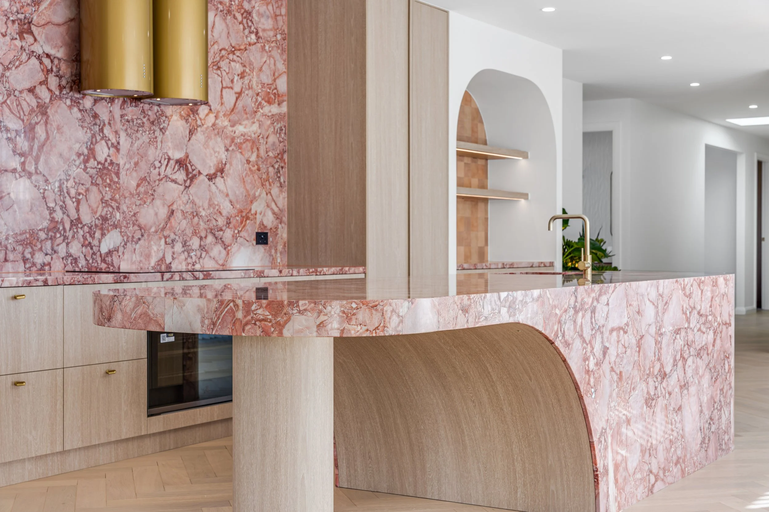 Modern kitchen with pink marble countertops and backsplash, light wood cabinets, and a gold faucet.
