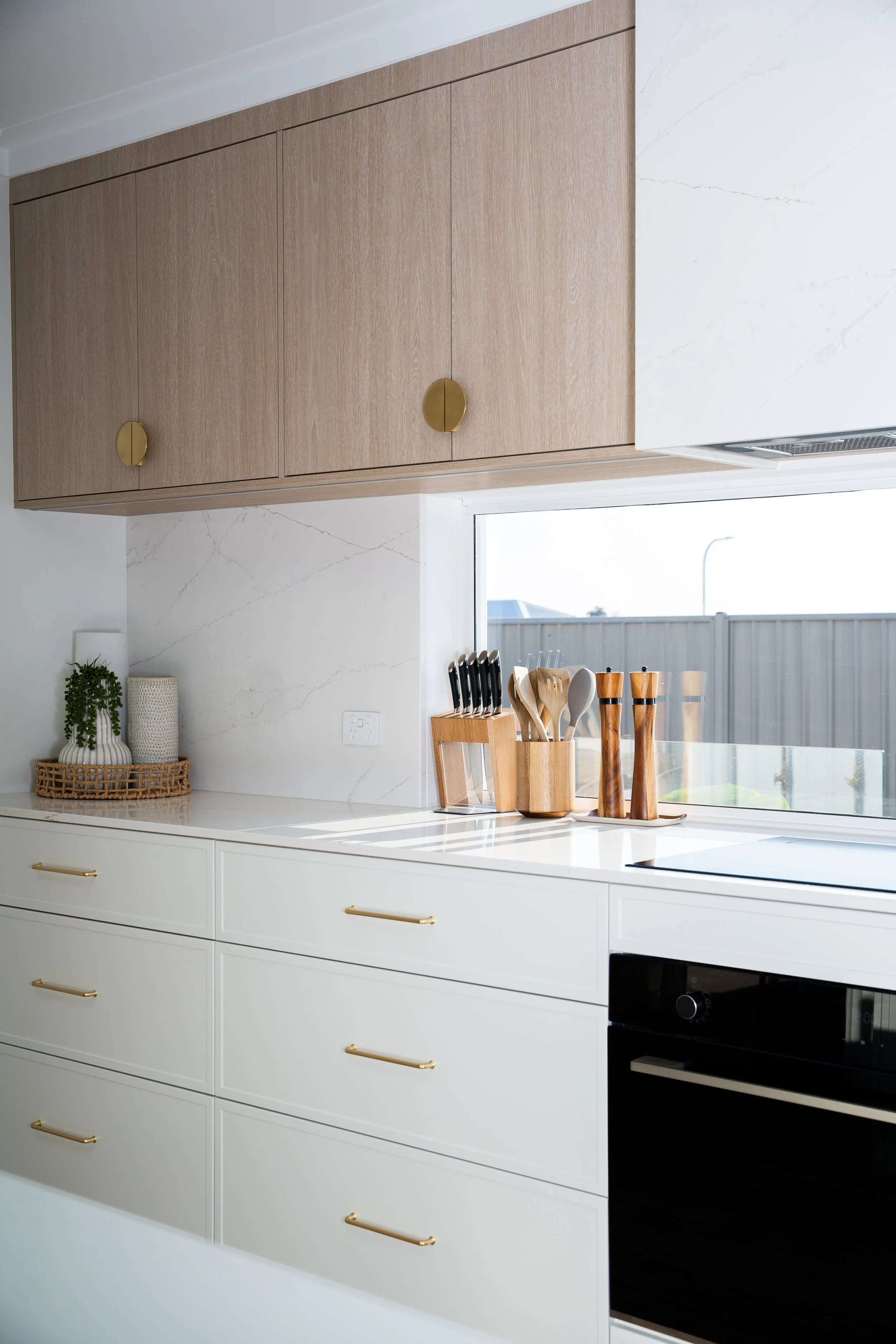Modern kitchen with white cabinets, gold handles, a marble backsplash, and wooden utensils in a holder on the countertop. There are plants and a window nearby.
