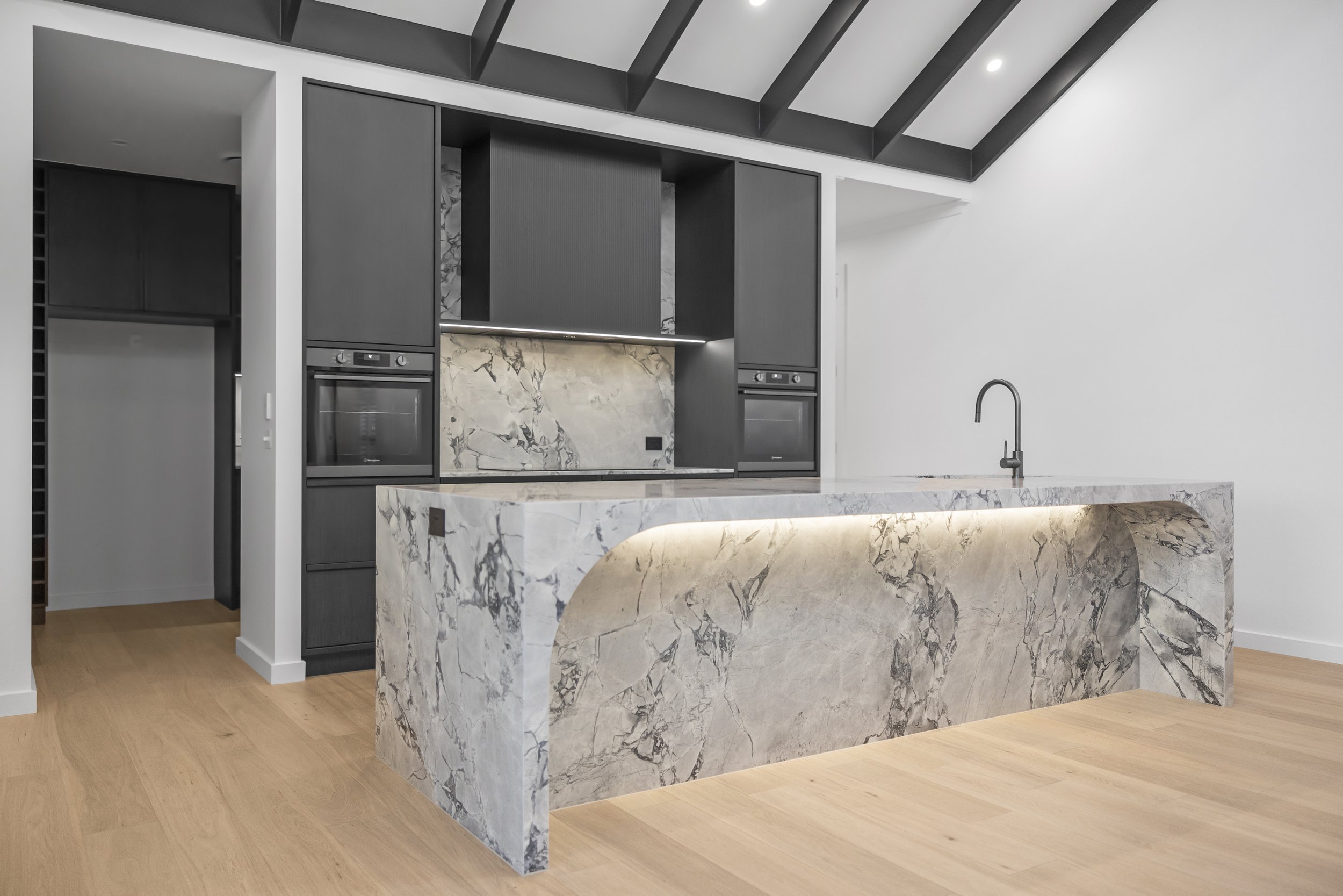 Modern kitchen with black cabinets, marble island, and a marble backsplash, with light wood flooring and a sloped ceiling with dark beams.