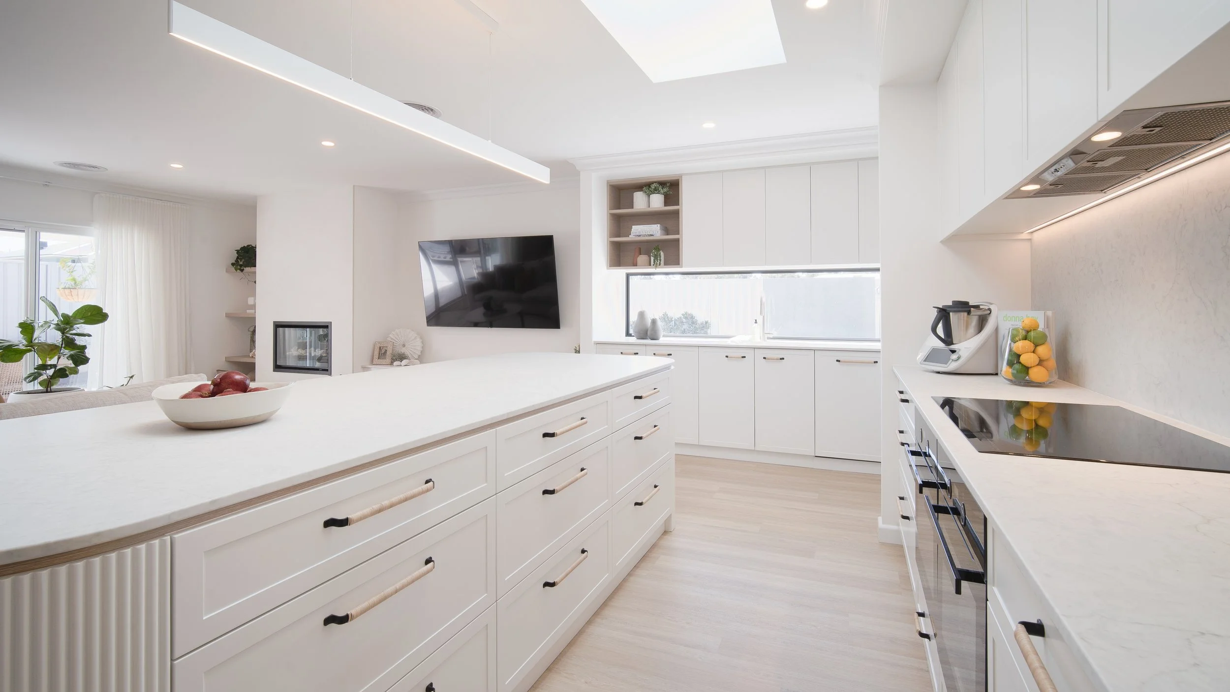 Modern white kitchen with a kitchen island, built-in appliances, and decor including a bowl of red apples, a plant, and vases, with natural light coming through a large window.