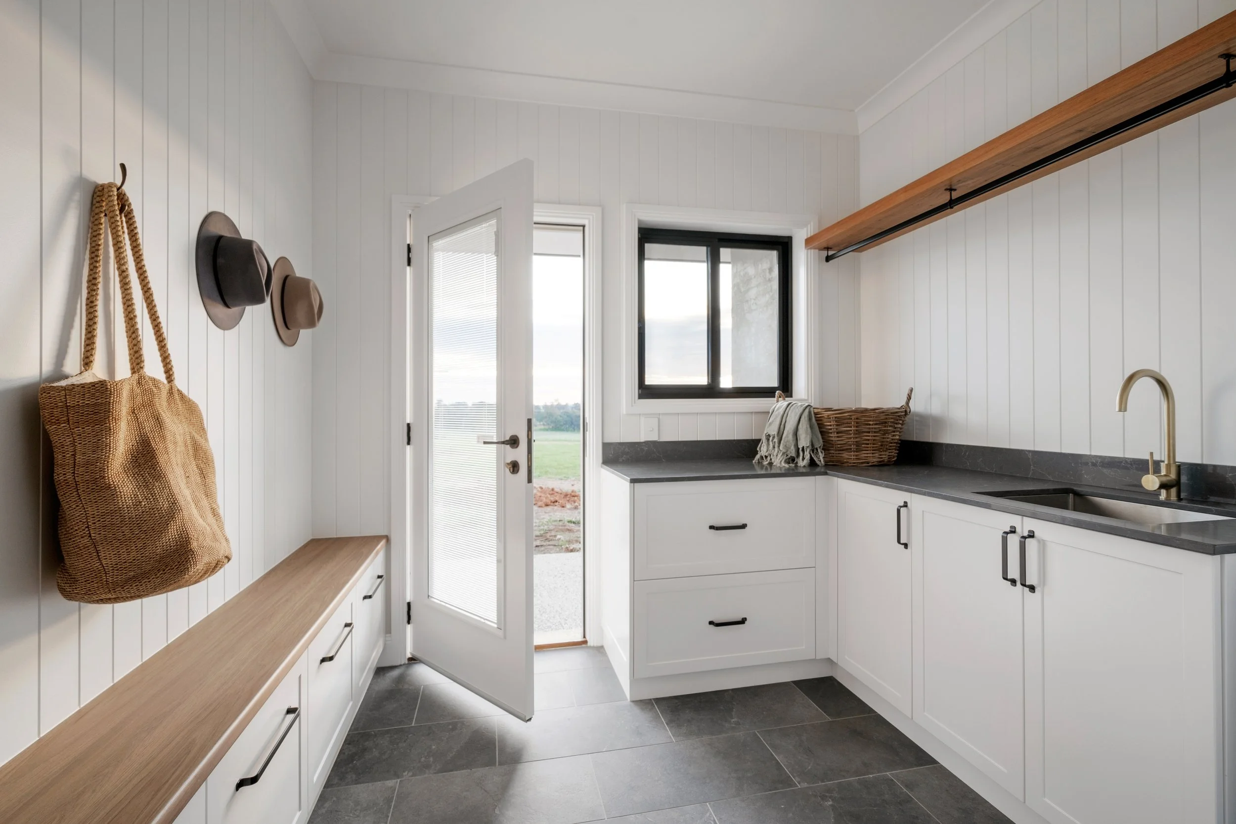 Modern kitchen with white shiplap walls, black accents, and a black countertop, featuring a window, open door, hanging hats, woven bag, basket, and a bench.