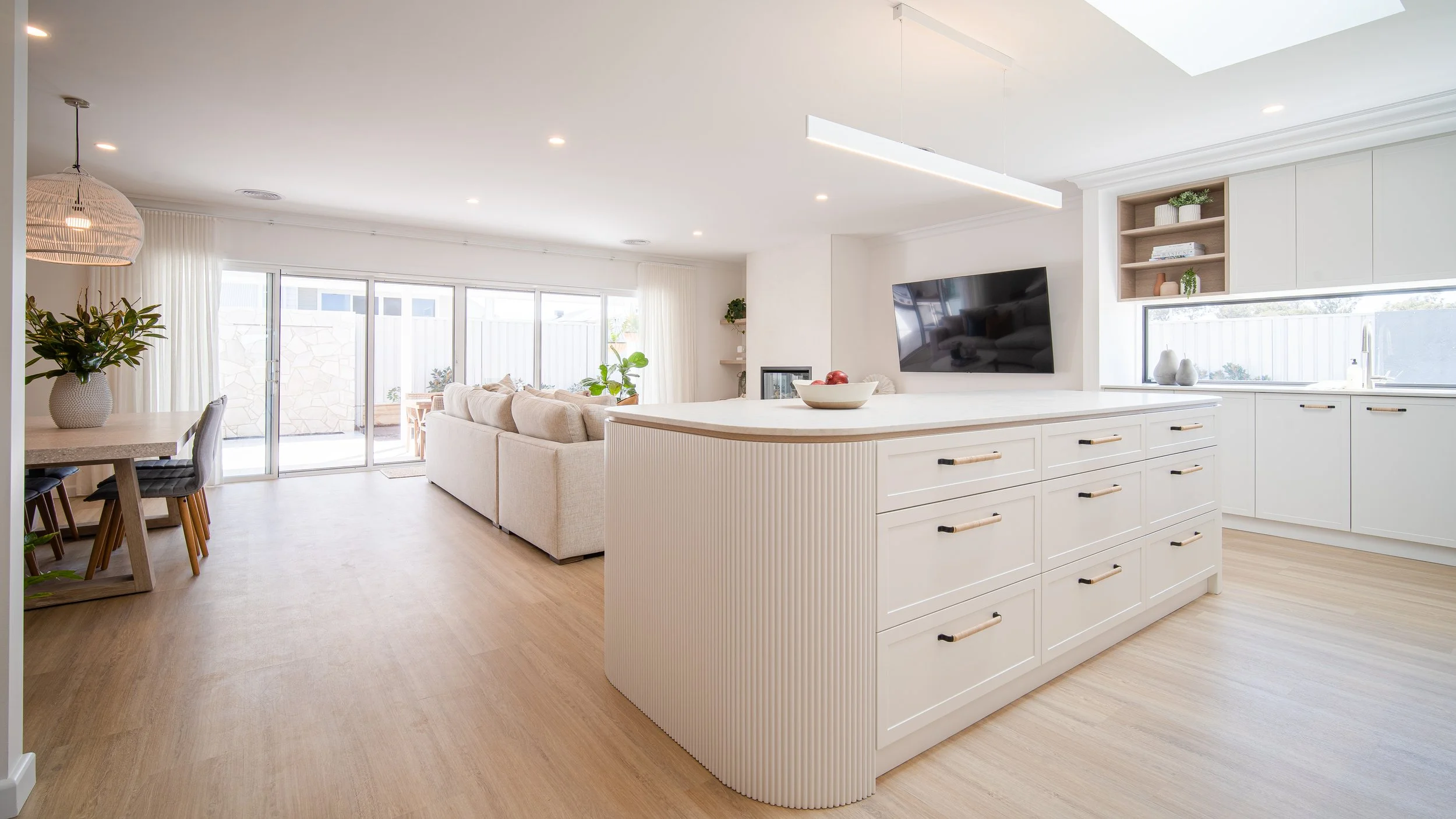 Bright, modern open-concept living room and kitchen with white cabinetry, a large island, a wall-mounted TV, and sliding glass doors leading to an outdoor patio.