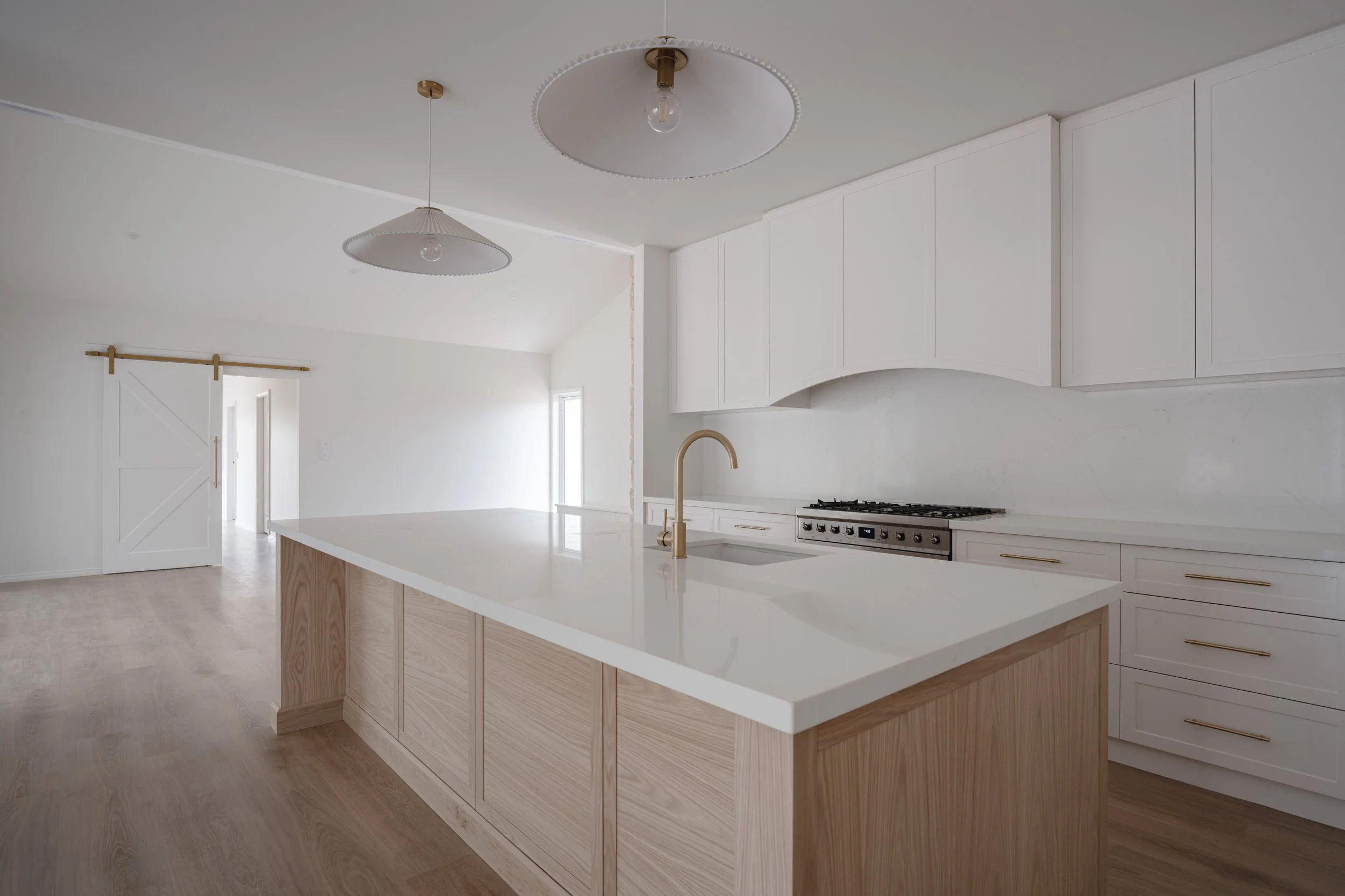 Modern white kitchen with a large island, gold fixtures, and light wood cabinets, with white walls and ceiling, and pendant lighting.