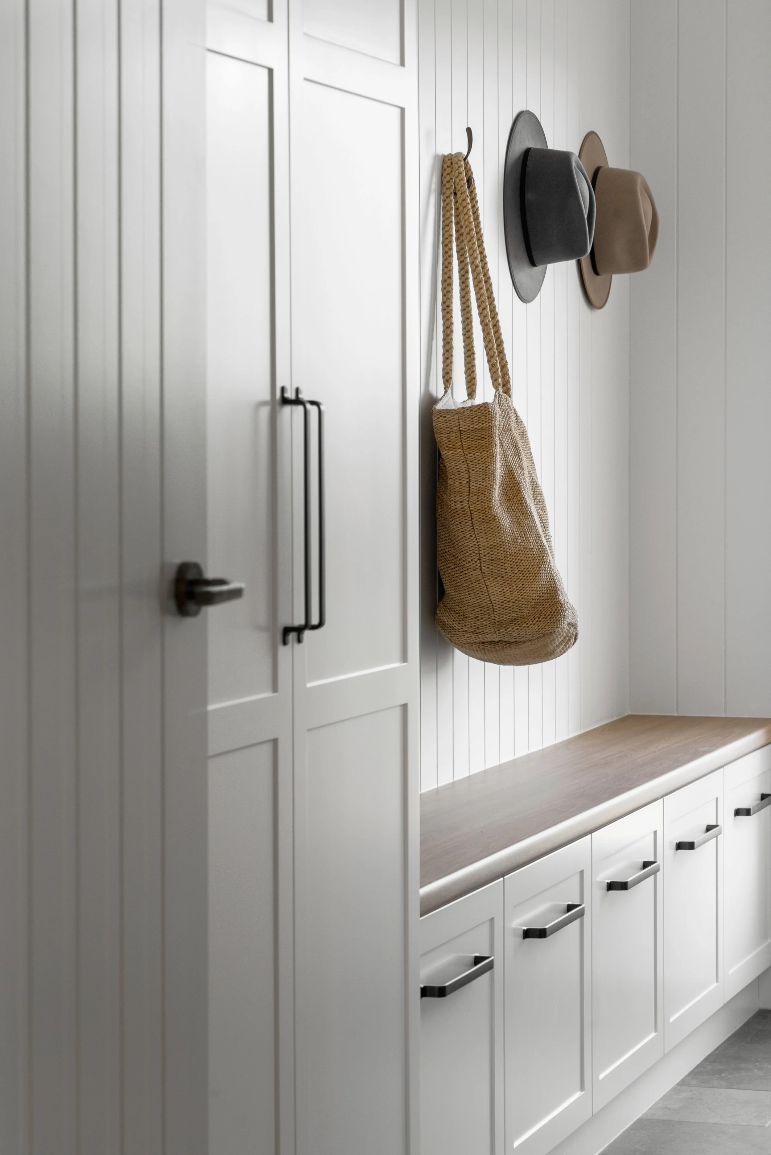 White kitchen cabinets with black handles and a wooden countertop. Above the countertop, a white paneled wall has a row of hooks holding two hats, a gray fedora and a beige hat, and a woven tote bag hanging from one hook.