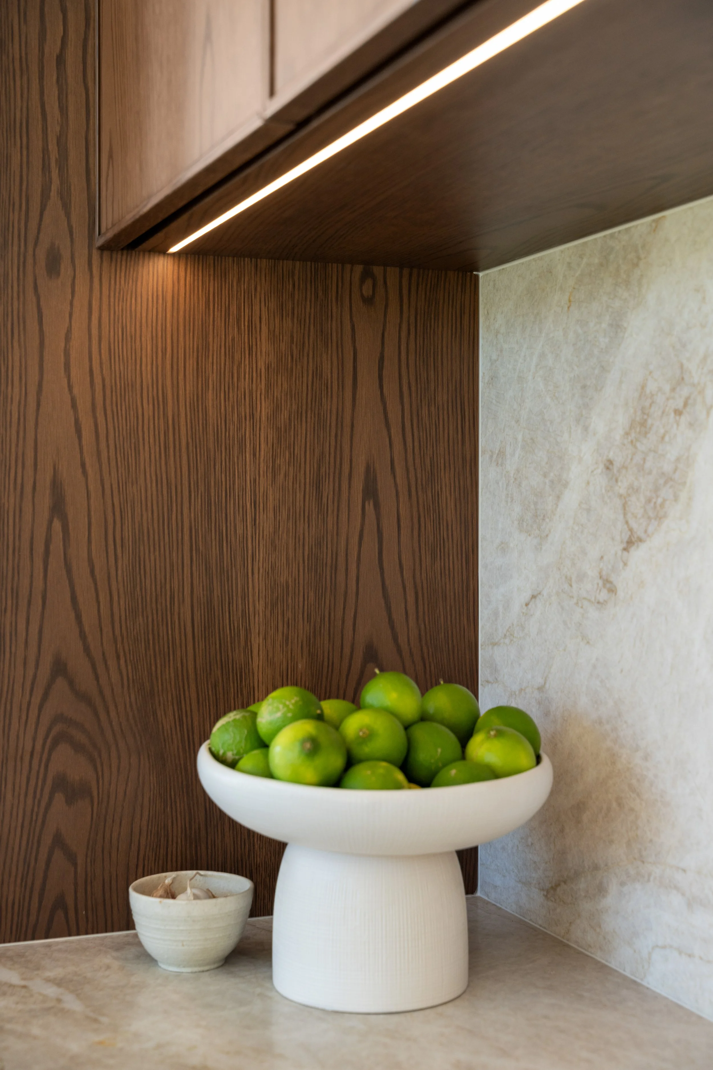 A white planter with a textured surface filled with green limes is placed on a beige countertop. Behind it, a wooden wall and a marble wall are visible. To the left, there is a small white bowl with garlic cloves.