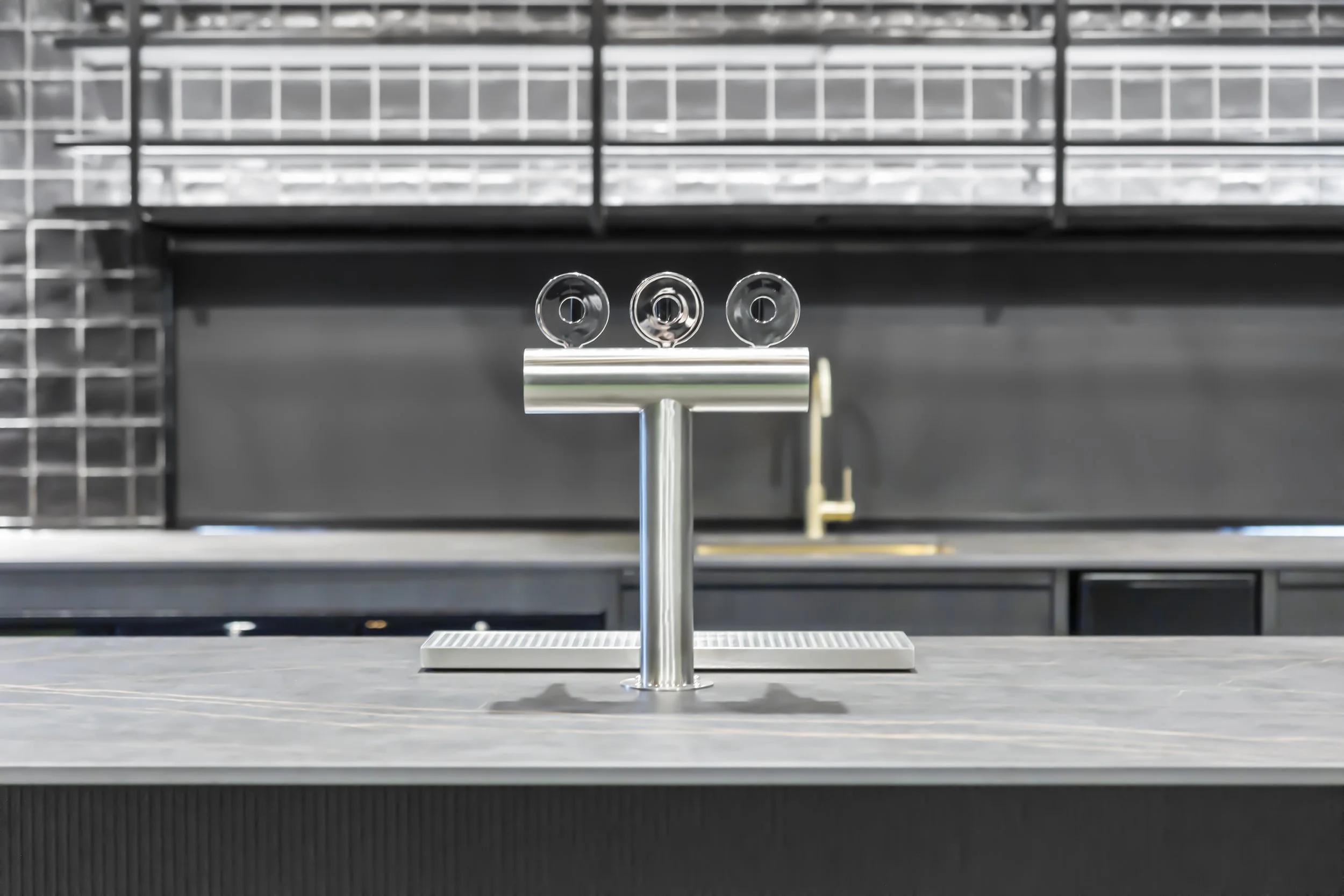 A stainless steel beverage dispenser with three spouts, set on a countertop in a commercial kitchen or cafeteria, with metal shelving in the background.
