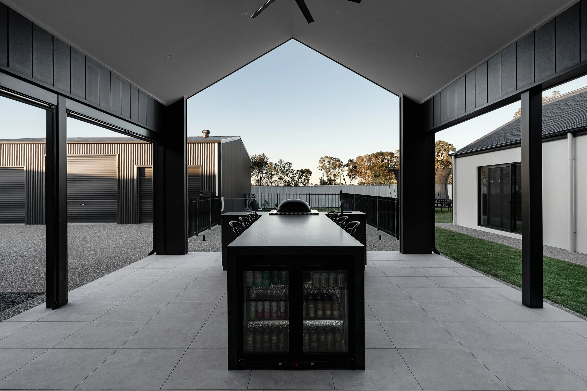 Modern outdoor patio with a large dining table, black chairs, and a built-in grill, covered by a ceiling with large opening to the outside yard featuring trees and neighboring buildings.