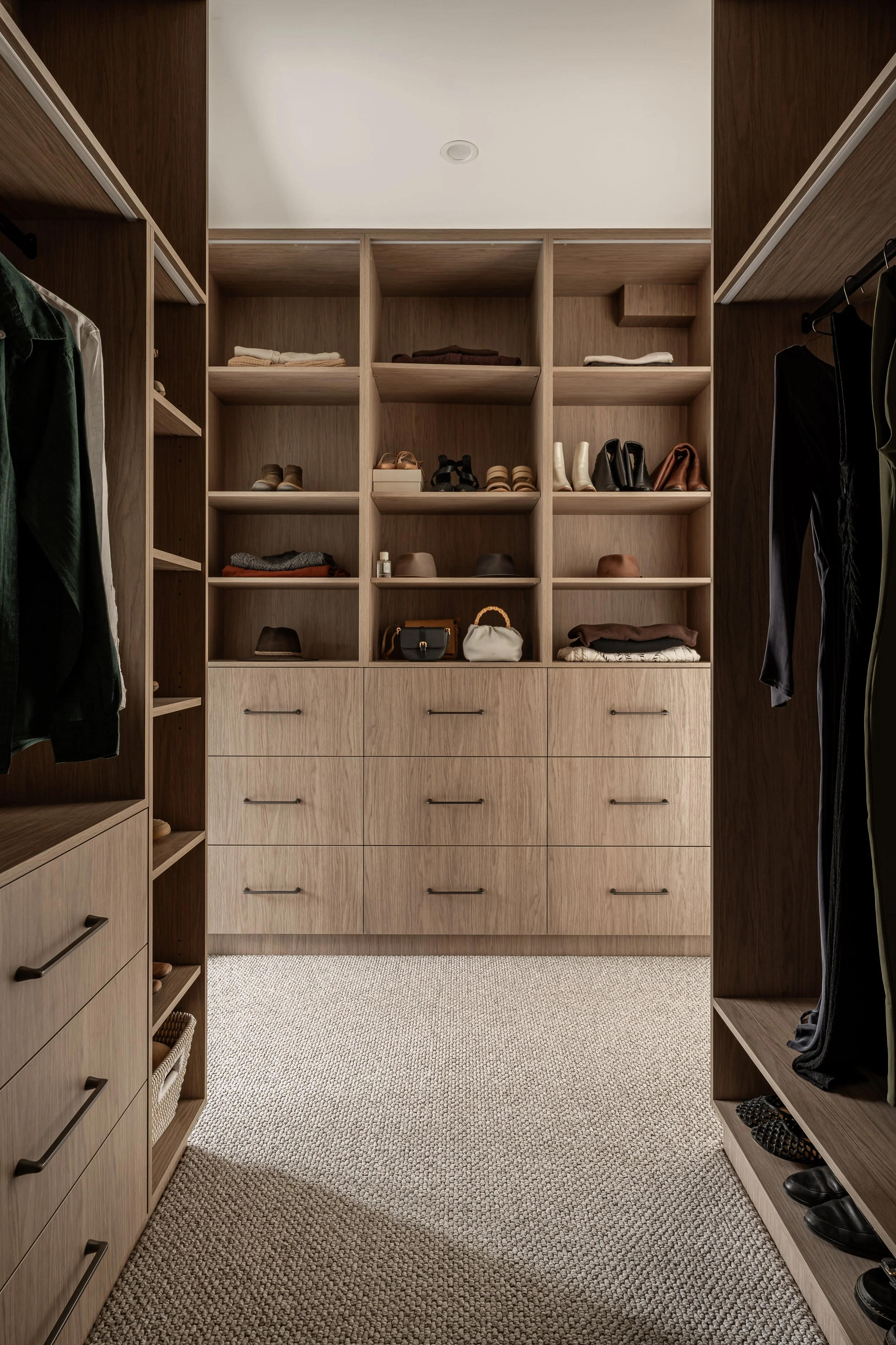 Walk-in closet with wooden shelves and drawers, containing folded clothes, shoes, handbags, and hats, with hanging clothes on sides and a beige carpeted floor.