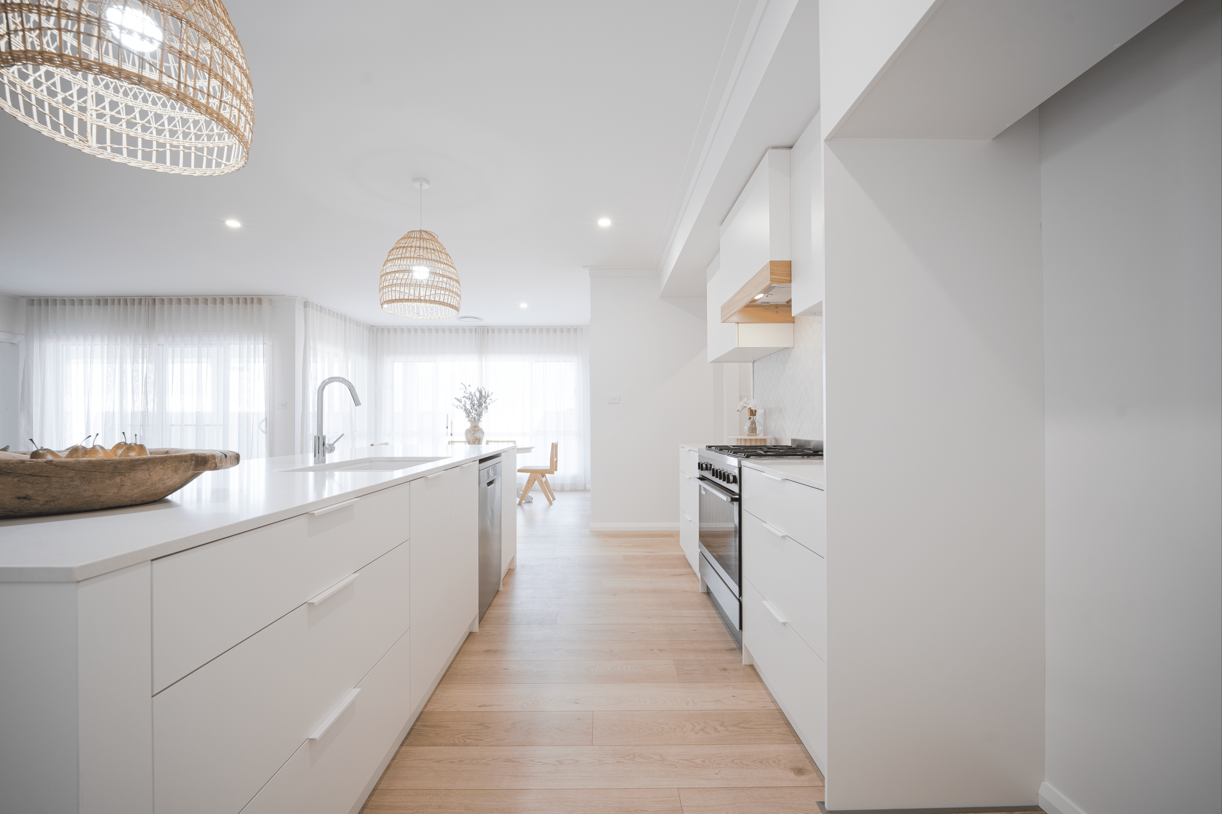 Bright, modern kitchen with white cabinetry, a wooden countertop, a stove, and a sink. Large windows with sheer curtains letting in natural light, and wicker pendant lights hanging from the ceiling.