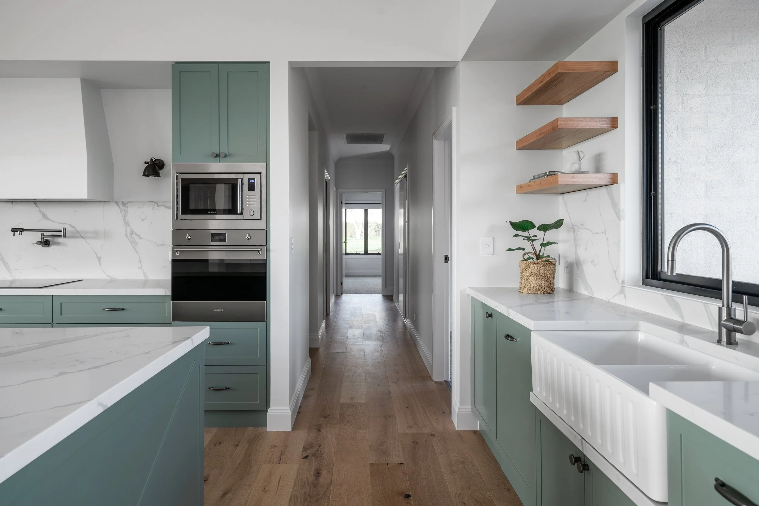 Modern kitchen with marble countertops, green cabinets, a microwave oven, open wooden shelves, a potted plant, and a window with a black frame.