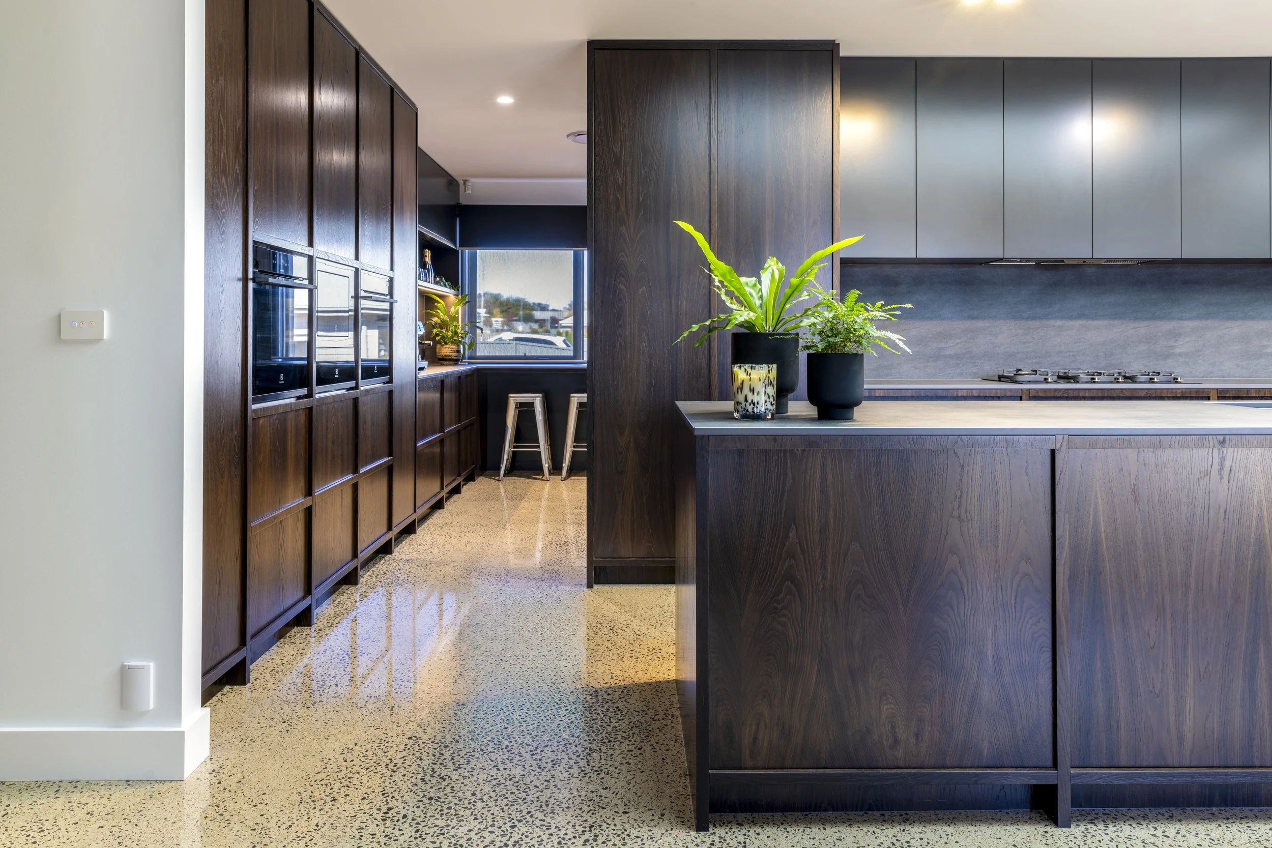 Interior view of a modern kitchen with dark wood cabinets, a blue backsplash, potted plants on a wooden kitchen island, and a window with two stools in the background.