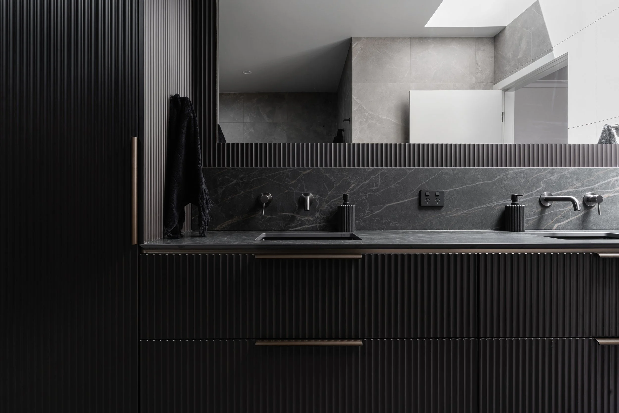 Modern bathroom with black vertically grooved door, gray marble countertop, black soap dispenser, black toothbrush holder, faucet, and a large mirror reflecting gray tiled walls and open door.