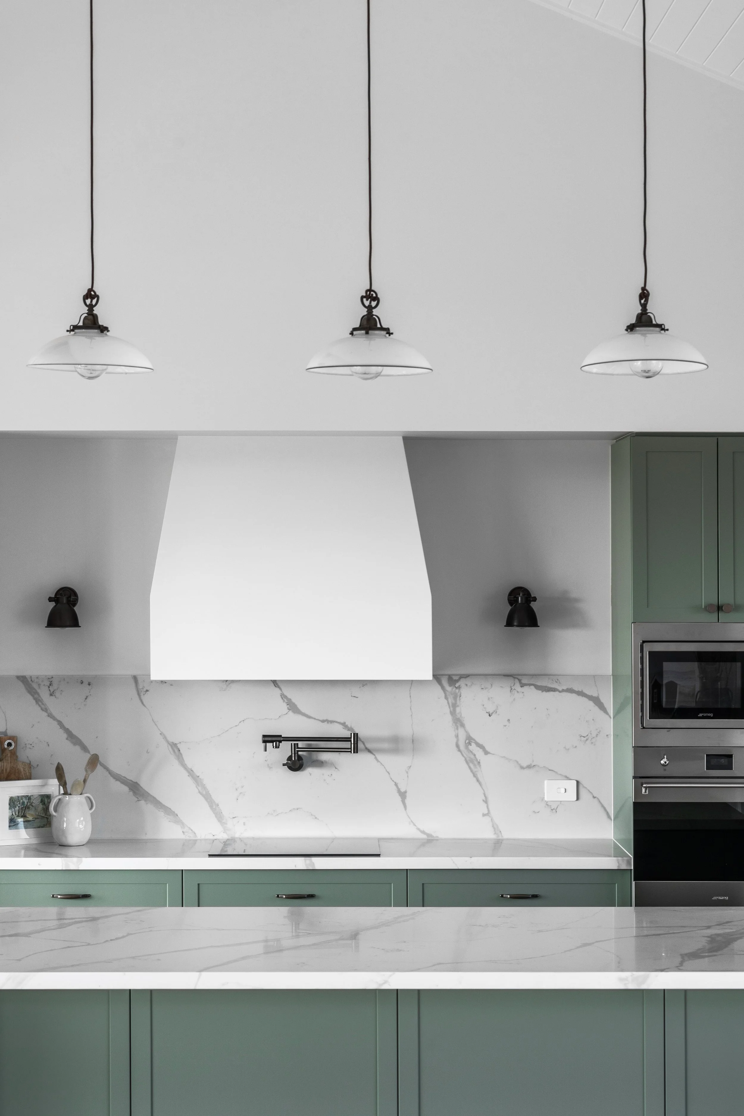 Modern kitchen with white marble backsplash, green cabinets, black pendant and wall lights, and metallic appliances.