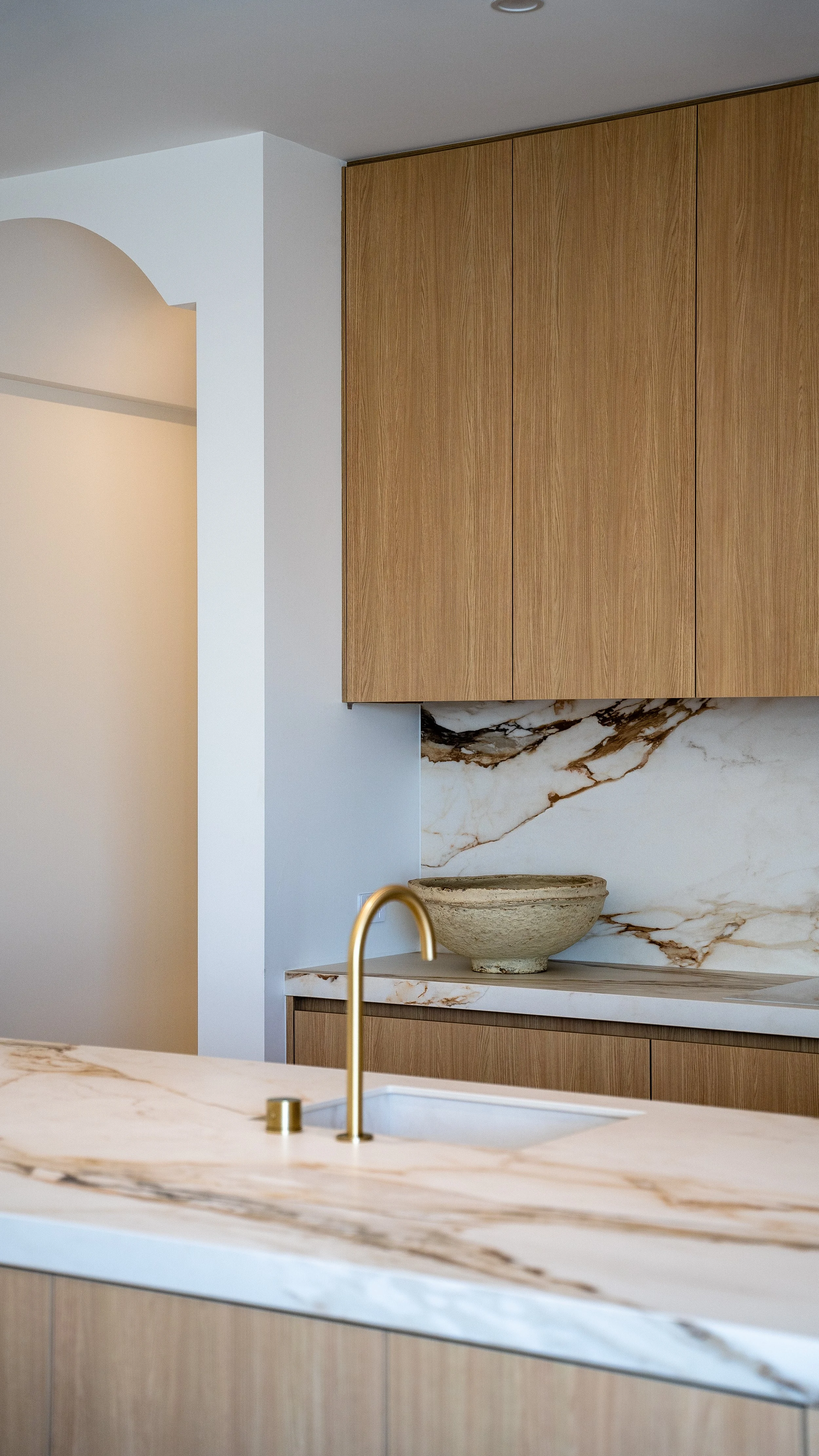 Modern kitchen with marble countertops, wooden cabinets, a beige bowl, and a gold faucet.