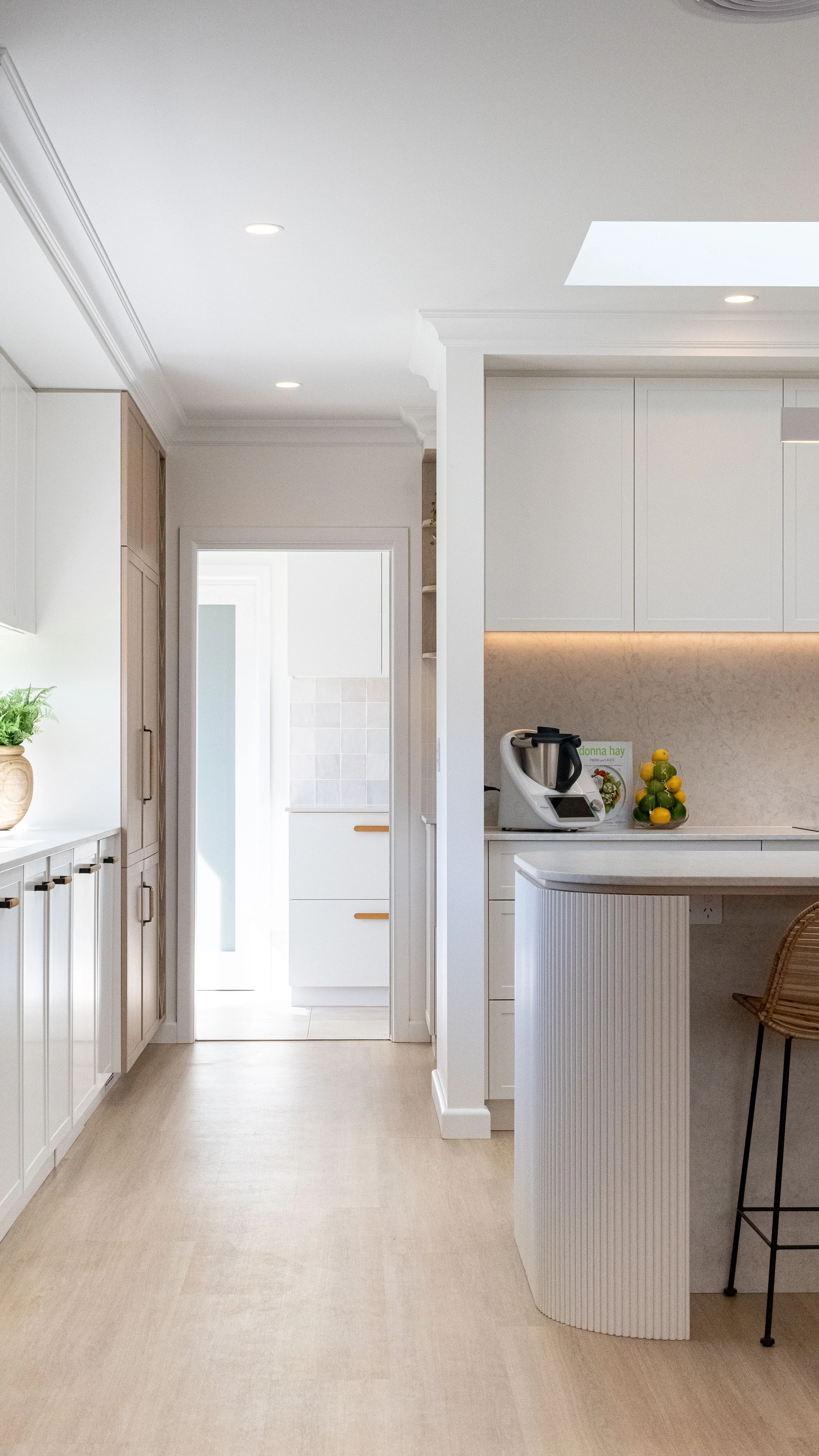Modern kitchen with white cabinetry, a countertop with a food processor, a decorative fruit sculpture, and a wicker chair.