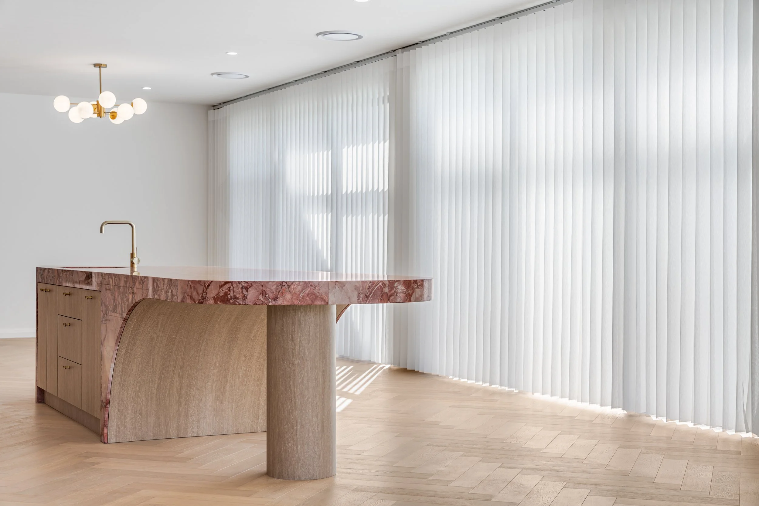 Modern kitchen with pink marble island, light wood cabinetry, white vertical blinds, hardwood floors, and a ceiling light fixture with multiple round bulbs.