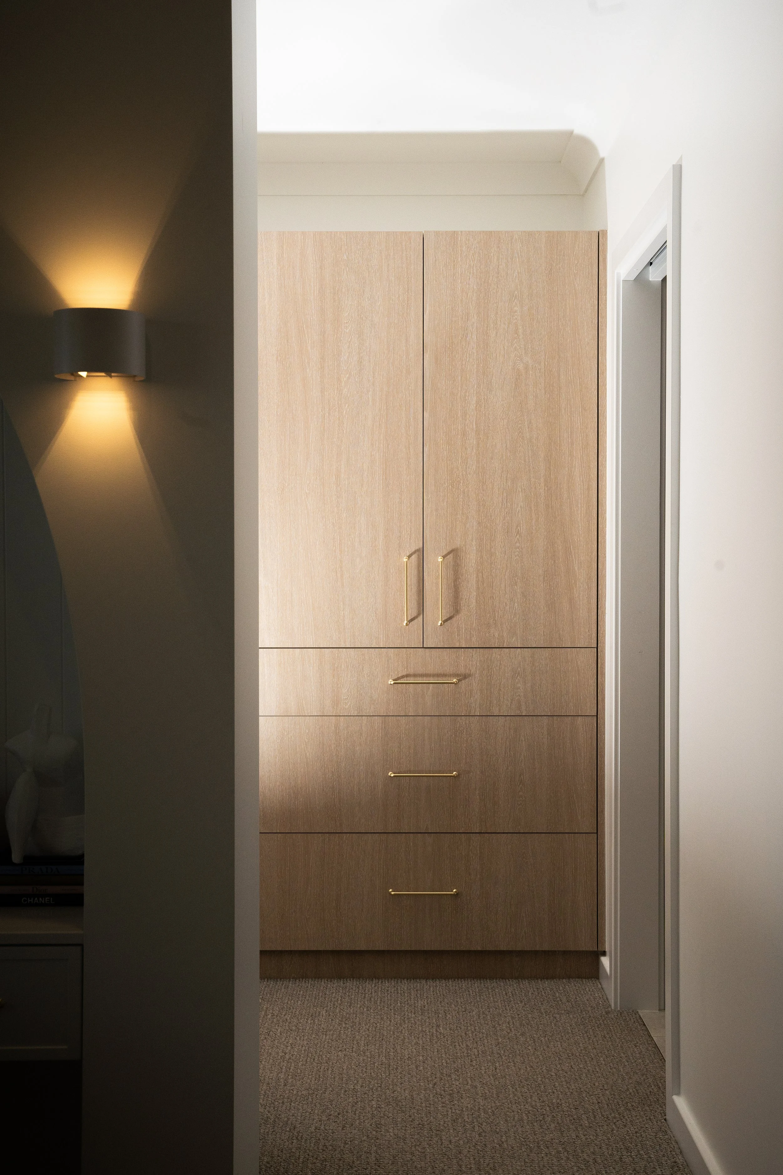 A closet with light brown wood doors and gold handles, situated in a room with beige carpeted flooring and white walls. There is a partial view of a dark-colored wall with a wall-mounted lamp casting light on the wall, creating an upward glow.