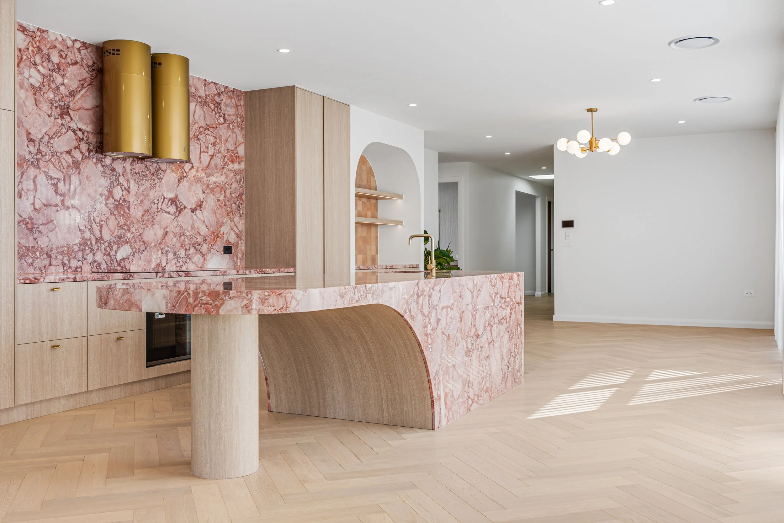 Modern kitchen with pink marble countertops and backsplash, light wood cabinetry, a gold faucet, and minimalist wood shelving, set in an open-concept space with white walls, light wood flooring, and ceiling lights