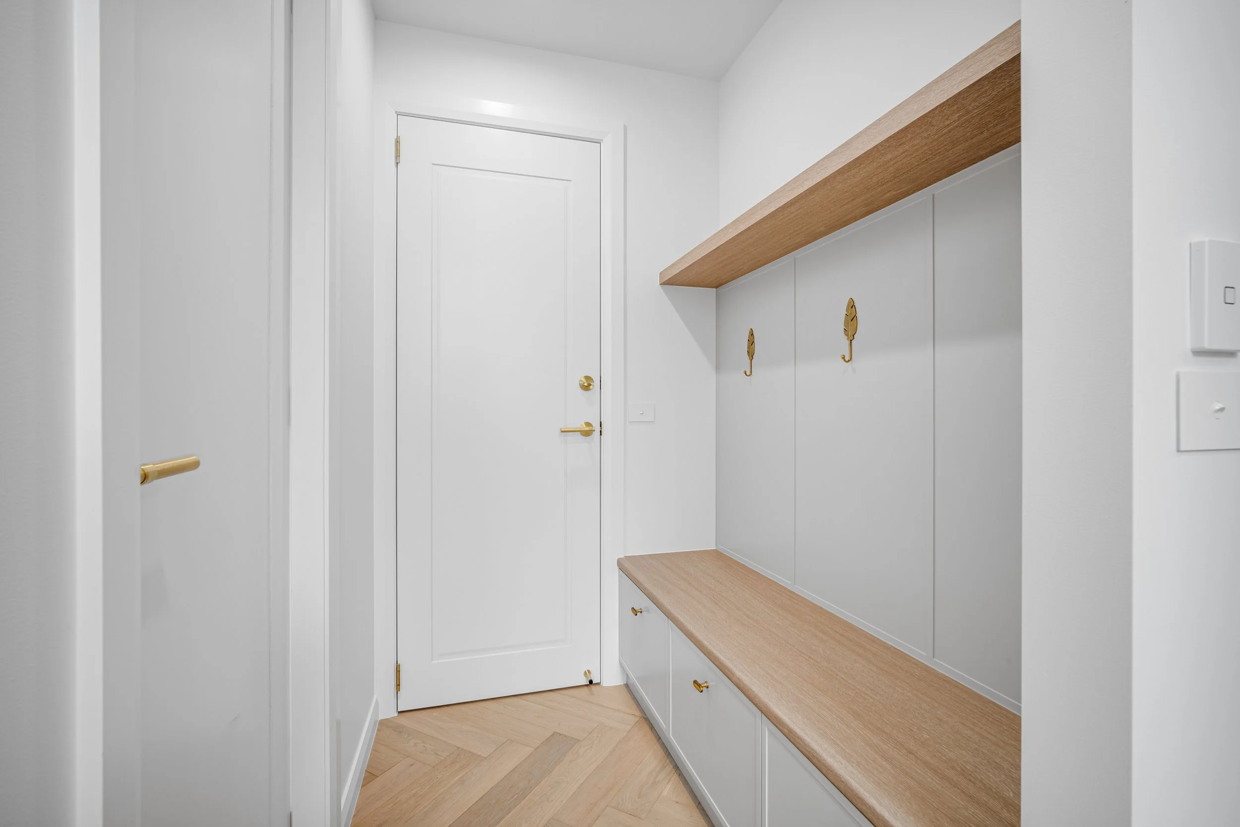 A modern entryway with white walls, a white door with gold hardware, a built-in wooden bench with drawers, and two decorative gold leaf hooks on the wall.
