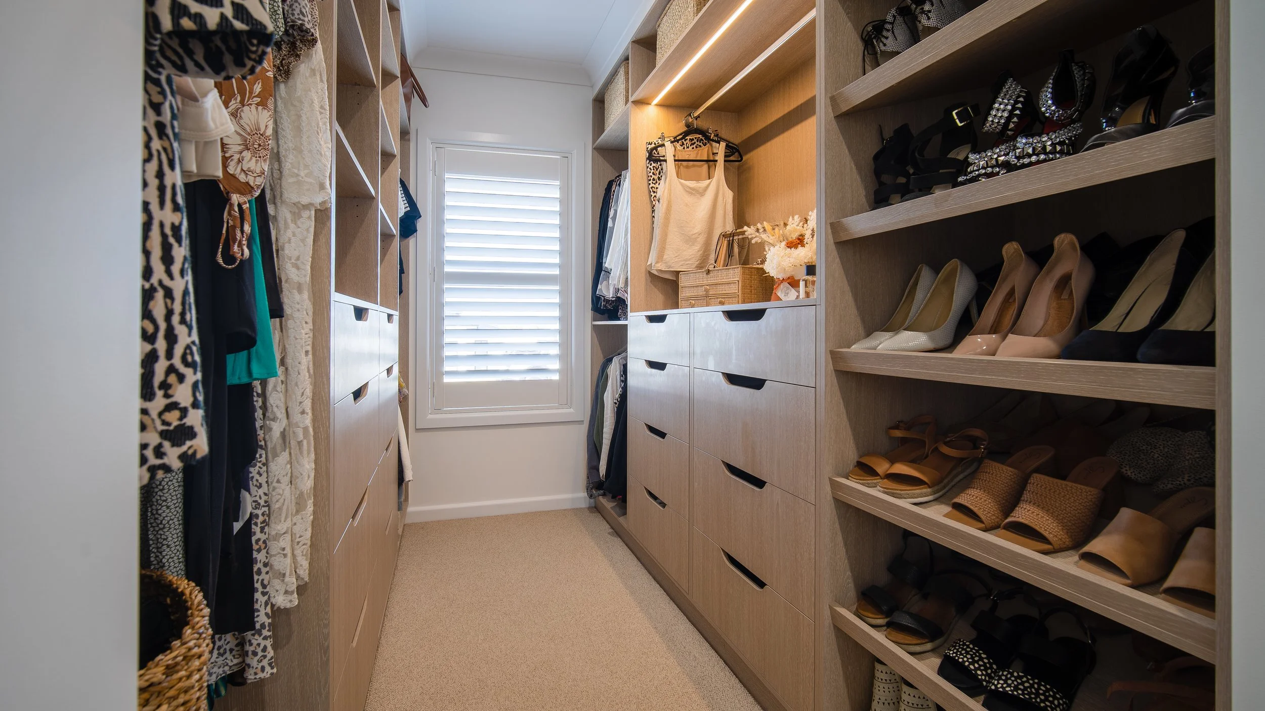 A spacious walk-in closet with wooden shelves and drawers, displaying clothes, shoes, and accessories. There is a window with blinds at the end of the room, allowing natural light to illuminate the space.