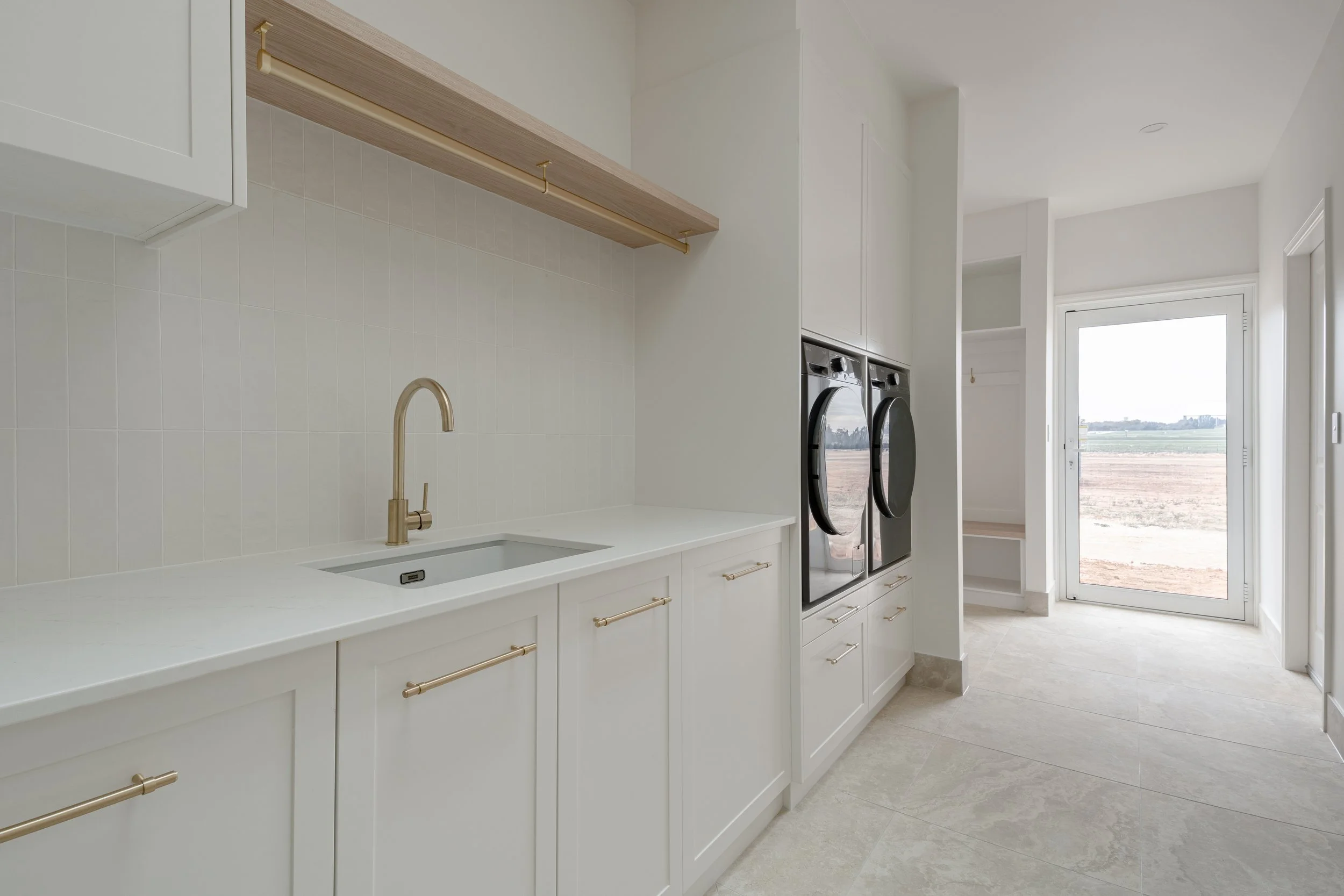 Clean, white laundry room with a sink, wooden shelves, and a washer and dryer, with a door leading outside to a rural landscape.