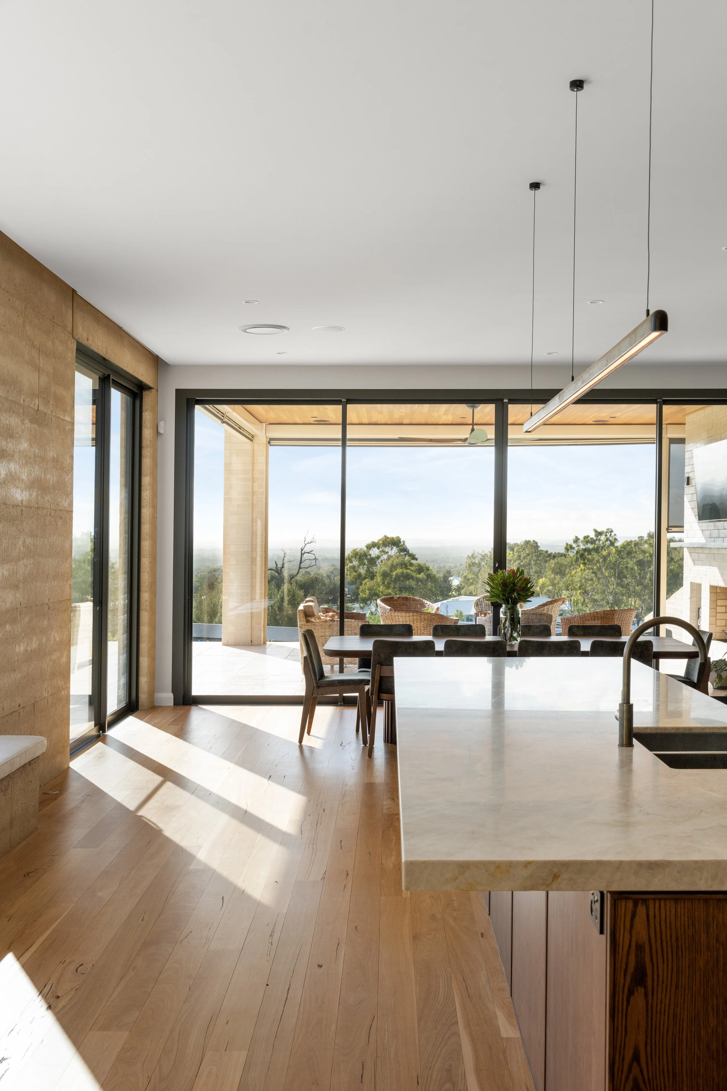 Modern dining area with wood flooring, a large white kitchen island, and a view of an outdoor patio with a dining table. Sunlight streams through large glass doors.