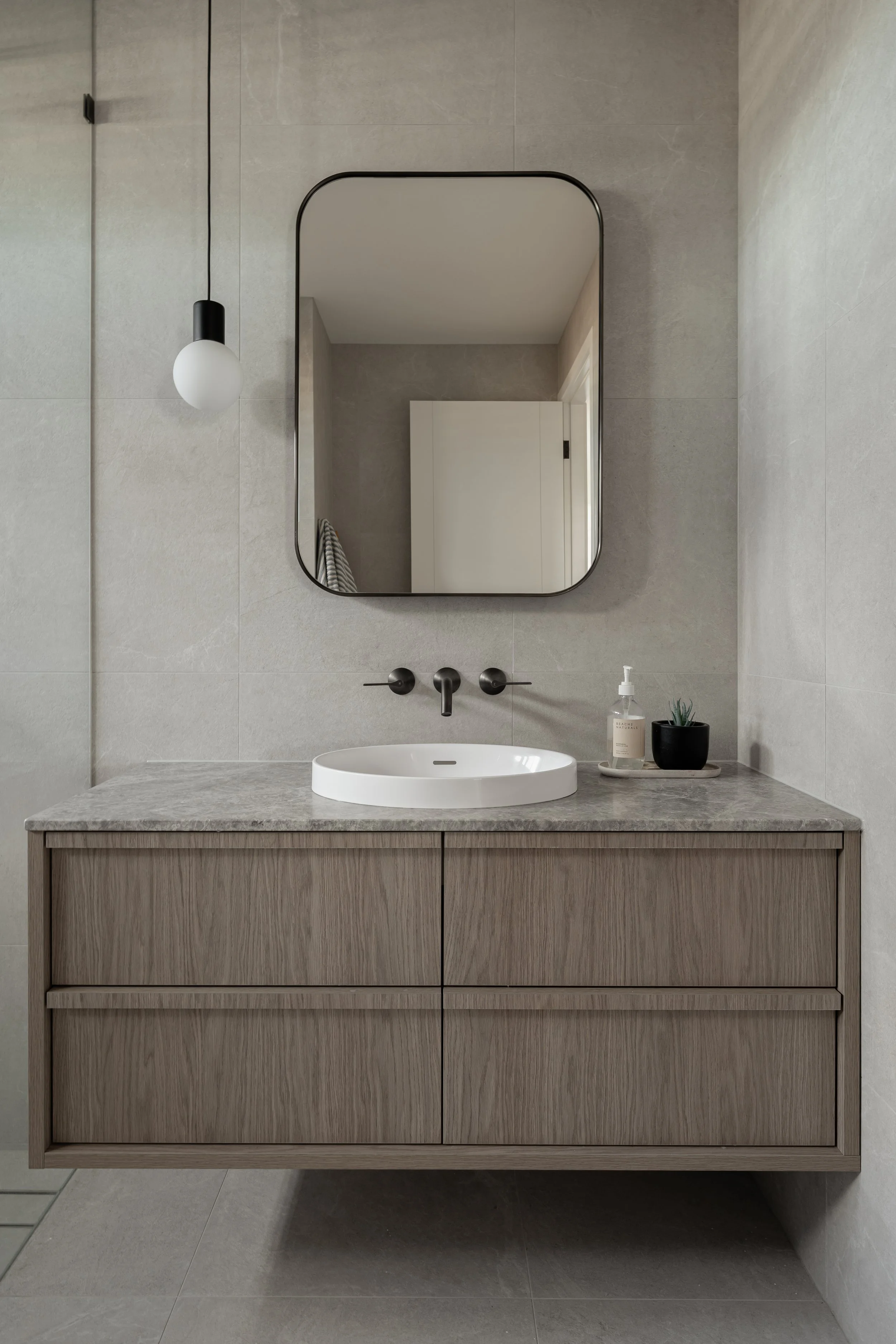 Modern bathroom vanity with a round vessel sink, a large rectangular mirror, a black hanging pendant light, and a soap dispenser with a plant on a small tray.