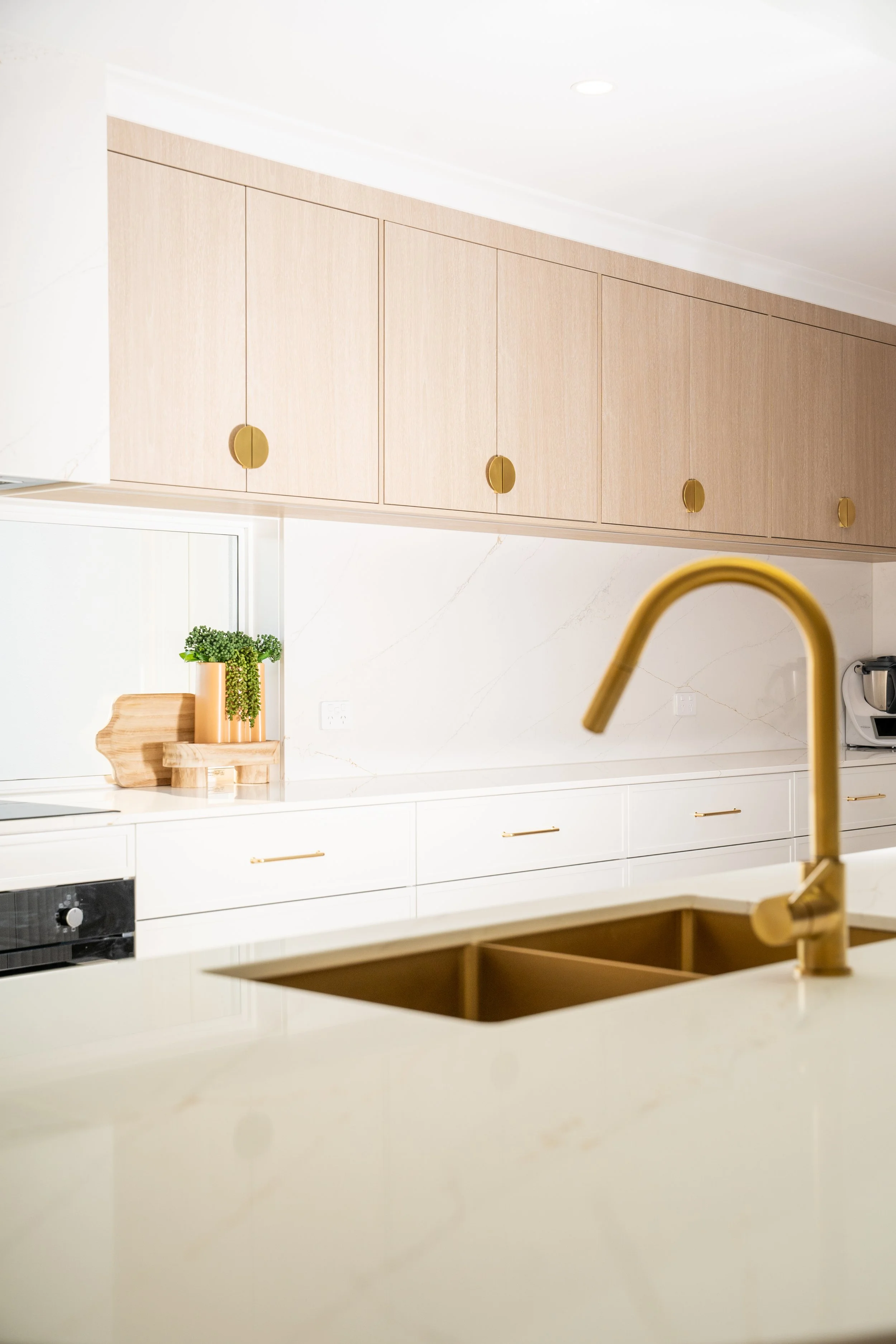 Modern kitchen with beige upper cabinets, white lower cabinets, a gold faucet, a sink, and a small potted plant on a cutting board on the counter.