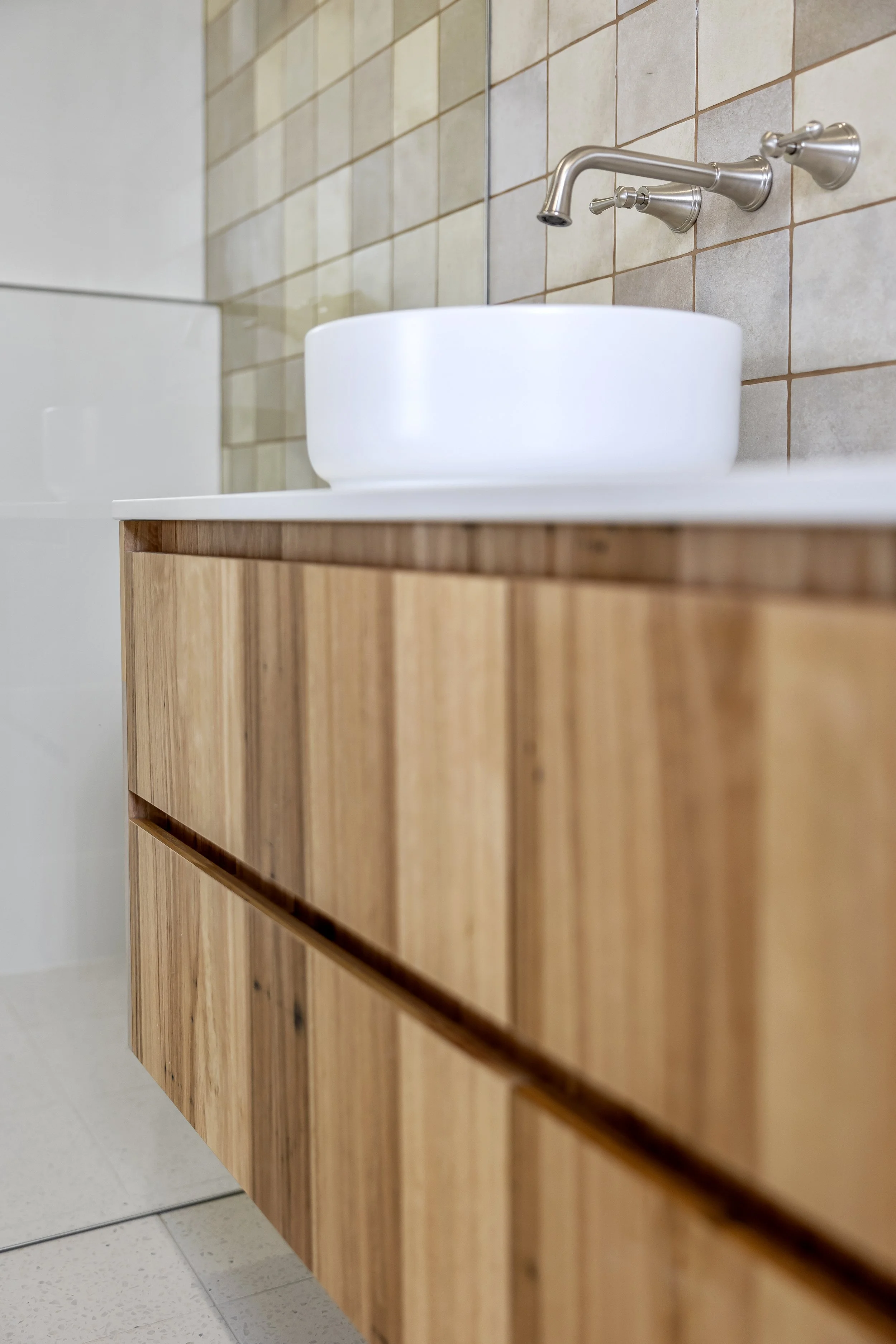 Close-up of a wooden bathroom vanity with a round white vessel sink and a wall-mounted brushed nickel faucet against a tiled wall.