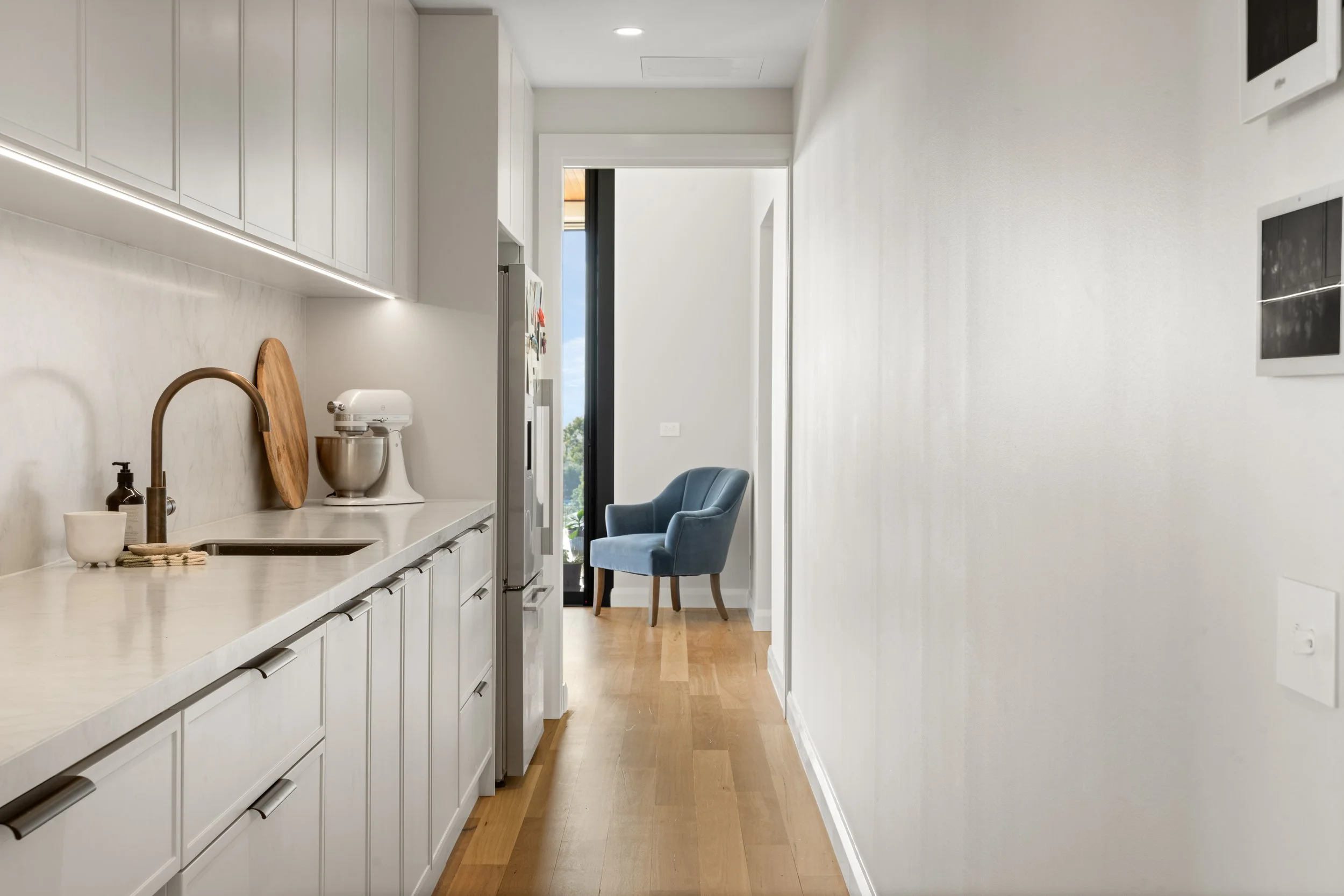 Modern kitchen with white cabinetry, marble countertop, and a sink with a bronze faucet. There is a white mixer on the counter and a wooden cutting board leaning against the wall. A small seating area with a blue upholstered chair is near the doorway