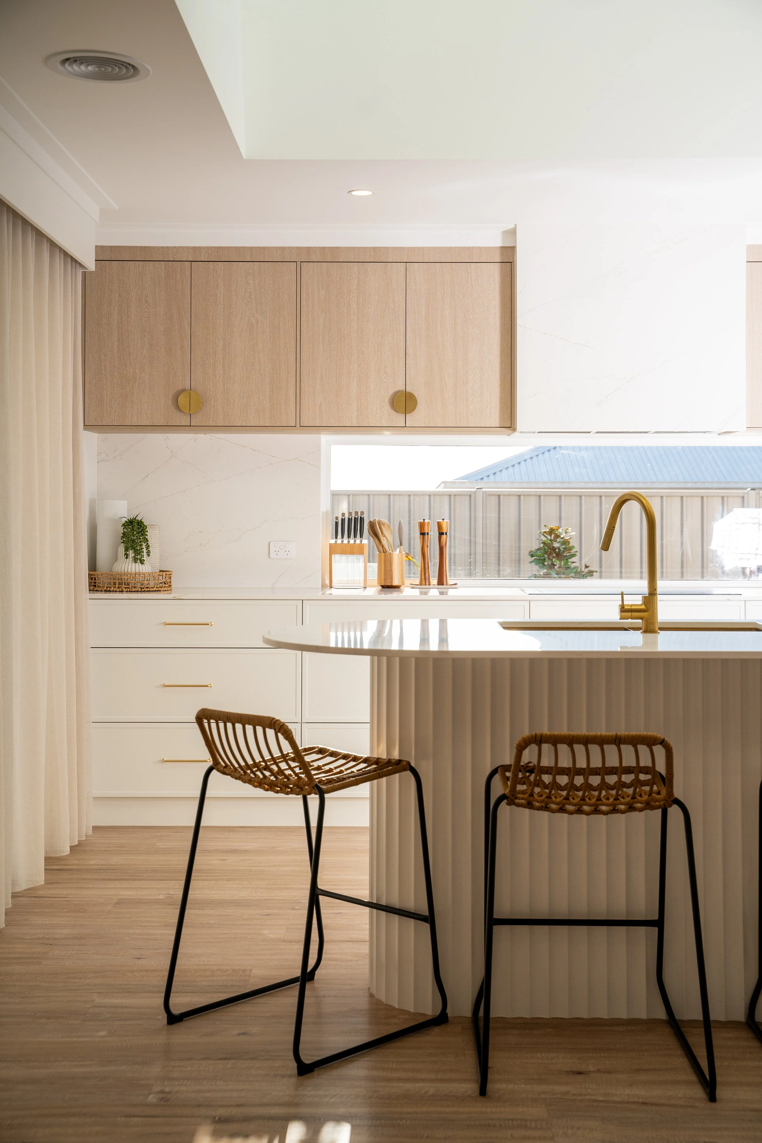 A modern kitchen with cream-colored cabinets, wooden accents, and a large window showing a blue roof outside. There are kitchen utensils on the counter, barstools at the island, and a brass faucet.