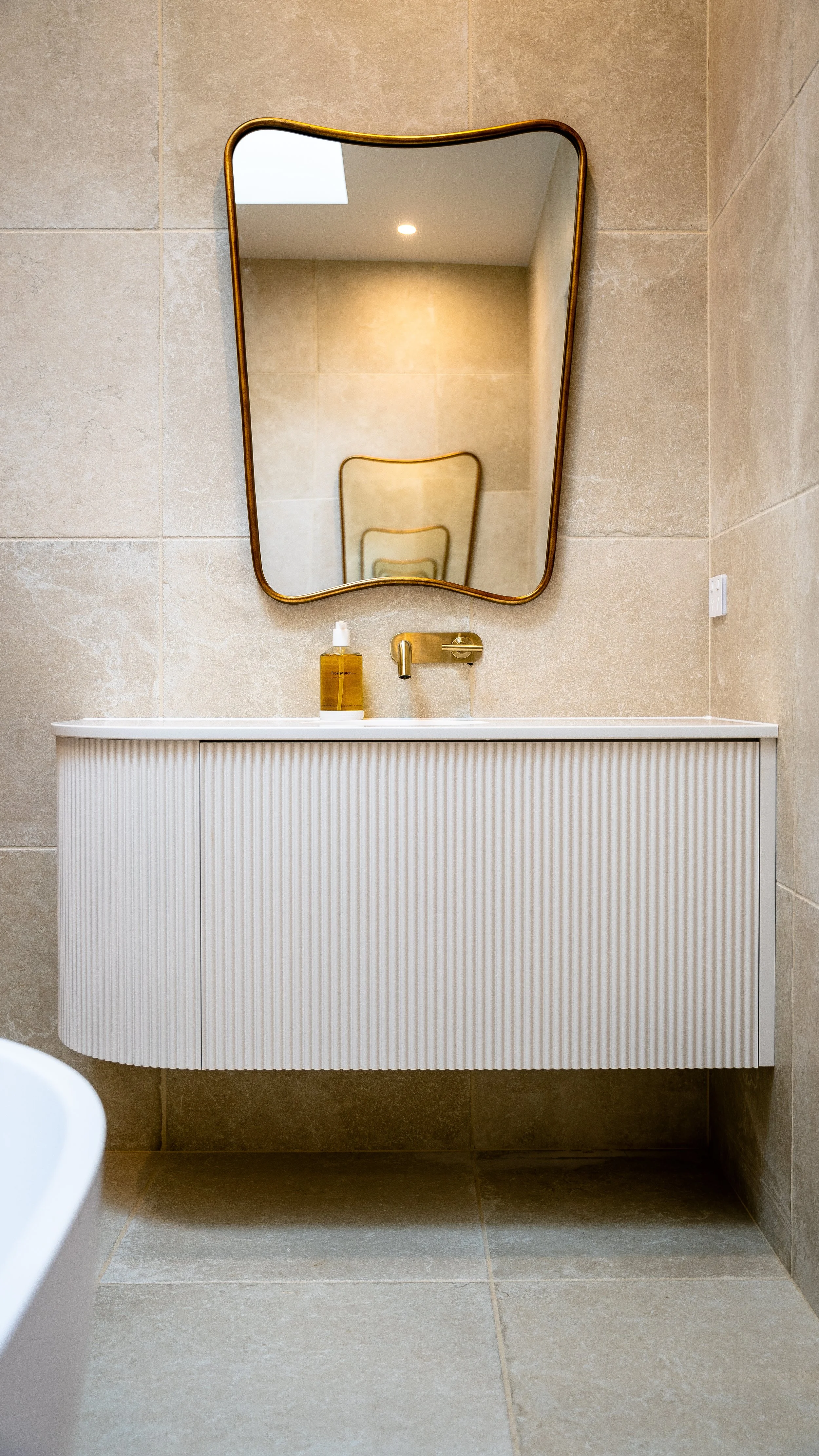 Bathroom with beige tiled walls and floor, white floating vanity with vertical grooves, gold wall-mounted faucet, rectangular mirror with gold frame, bottle of hand soap on sink, visible ceiling light and a skylight.