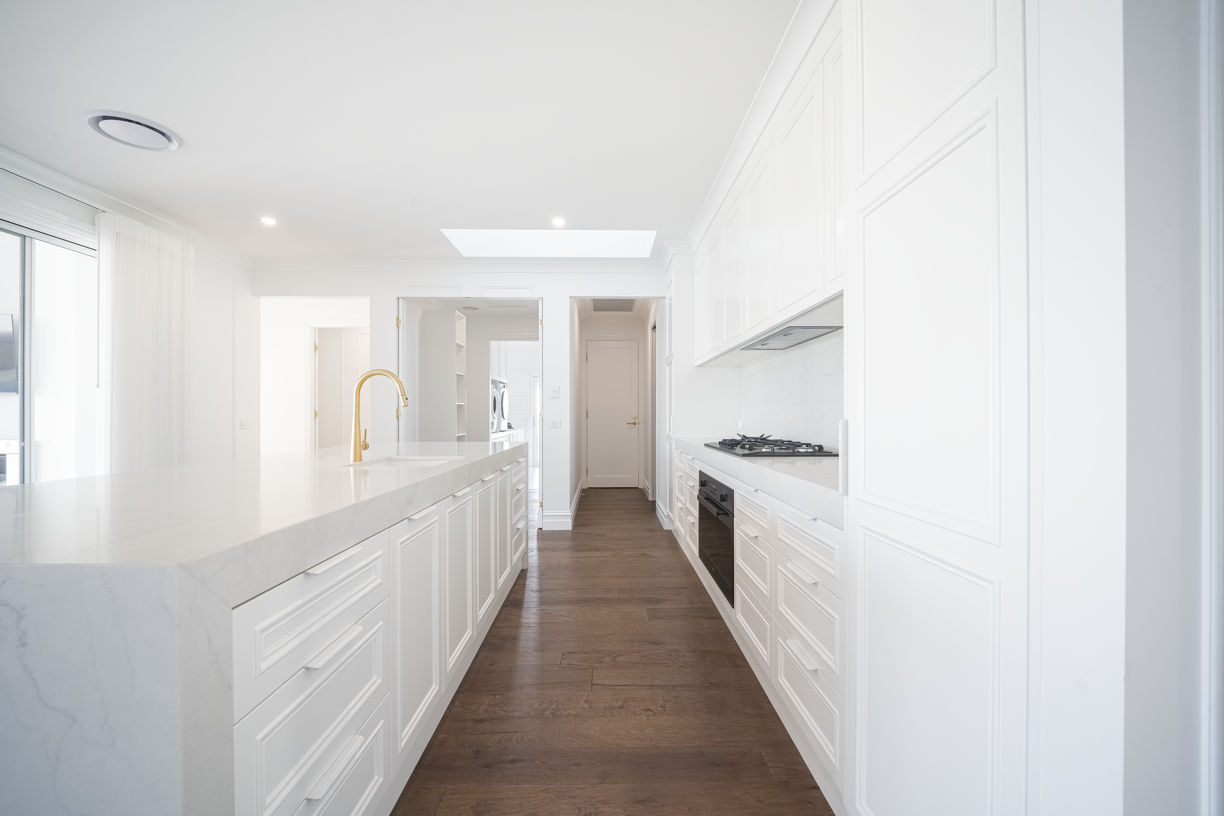 A modern kitchen with white cabinets, a white marble countertop, wooden flooring, and a gold-colored faucet. There is natural light coming in from a large window on the left.