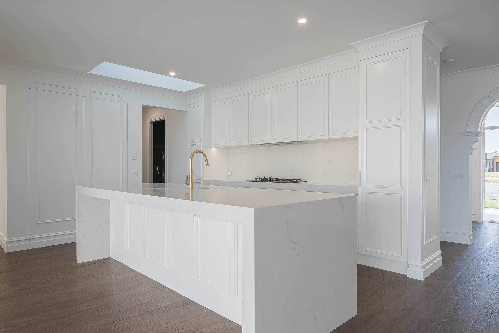 Modern white kitchen with island, marble countertops, gold faucet, and natural light from a skylight.