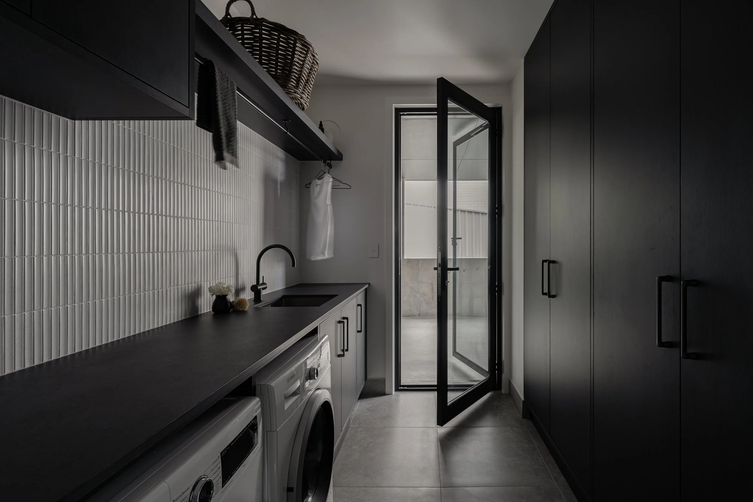 Modern laundry room with black and white color scheme, featuring black cabinets, a countertop, a white tiled backsplash, a washing machine, and an open door leading to an outdoor area with a concrete wall.