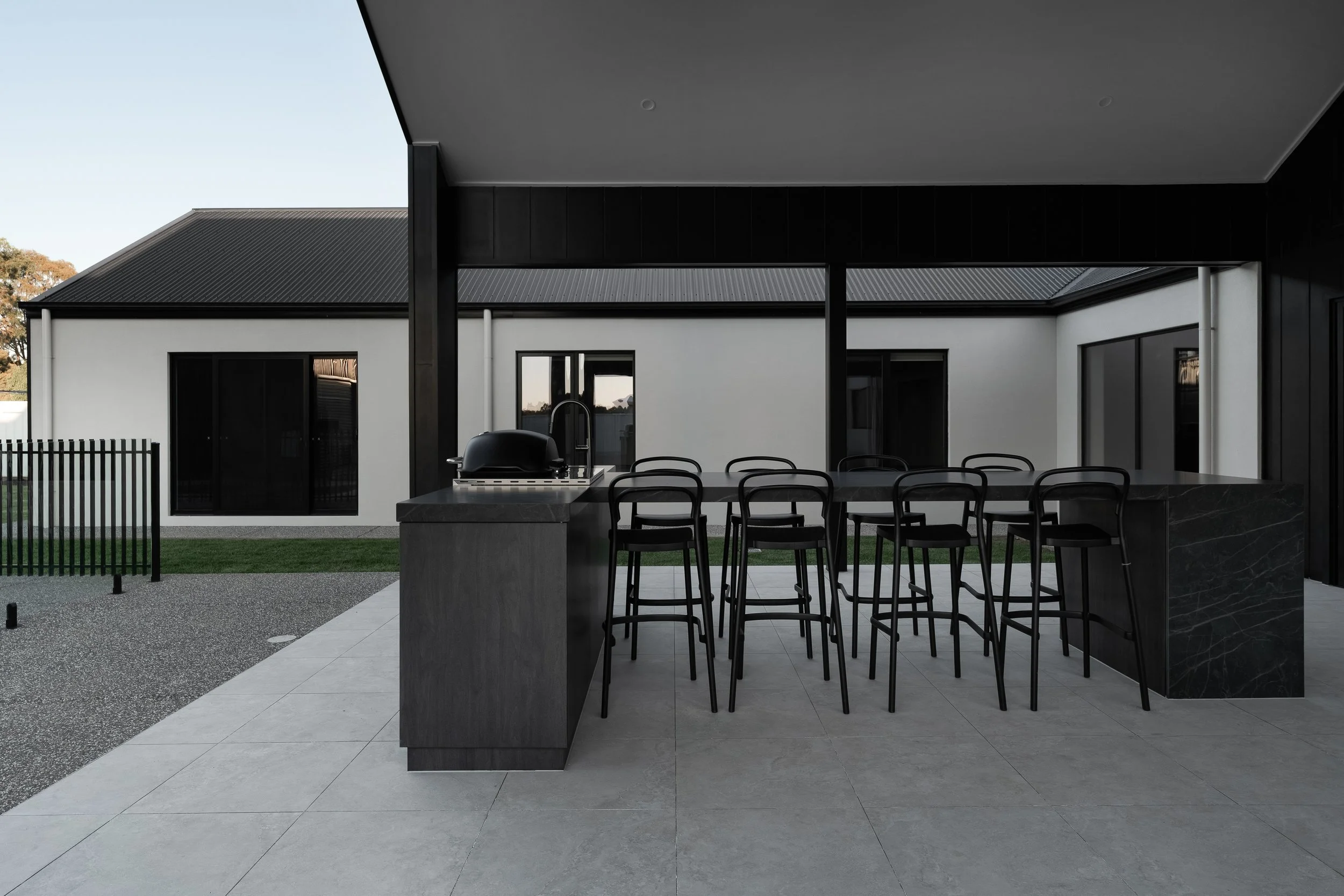 Modern outdoor patio area with a black dining table, black chairs, a built-in grill, and a view of a house with dark roof and white walls.